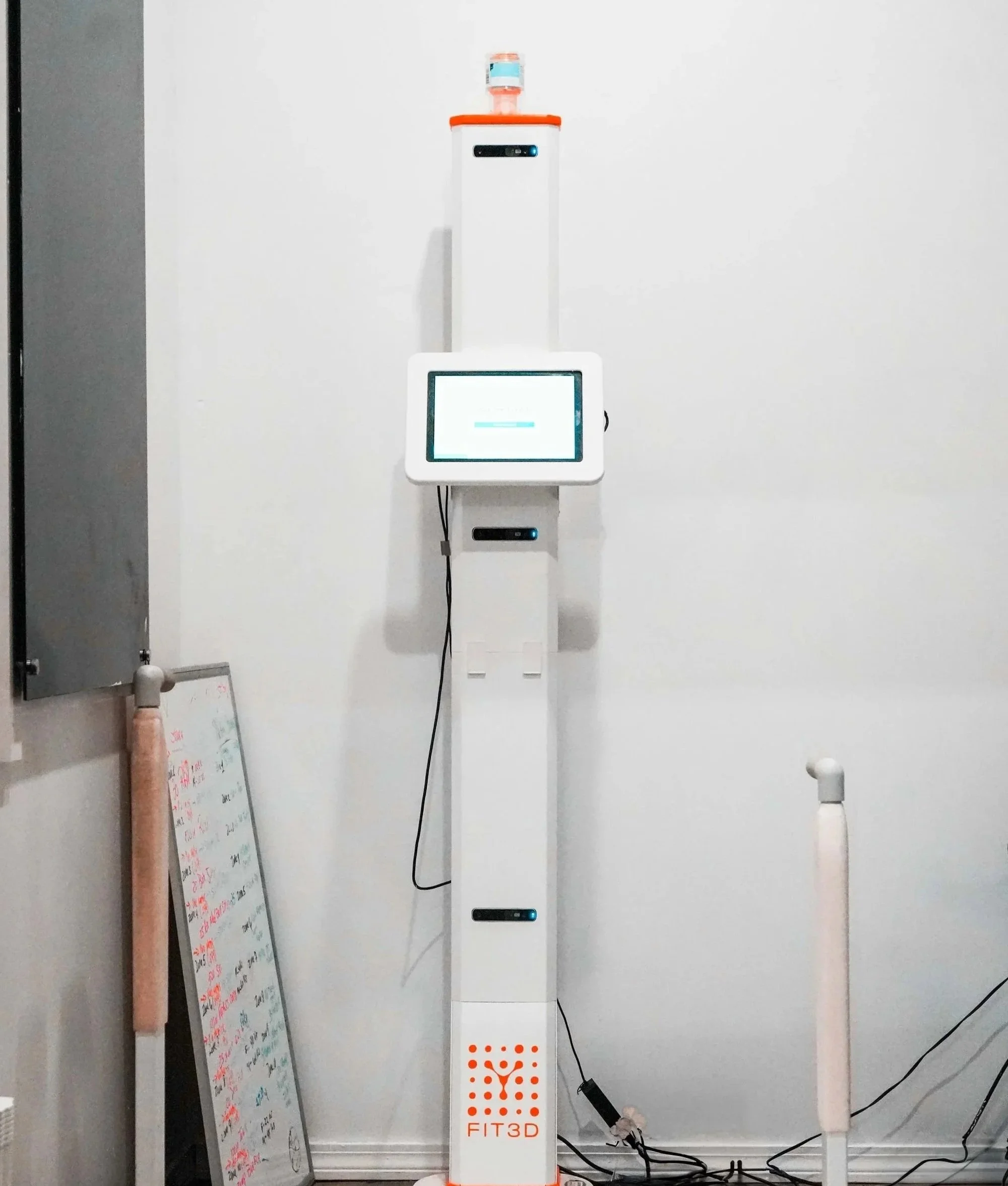 A tall white fitness device labeled 'FIT3D' with a touchscreen monitor and a small water dispenser on top, positioned against a plain white wall in a room.