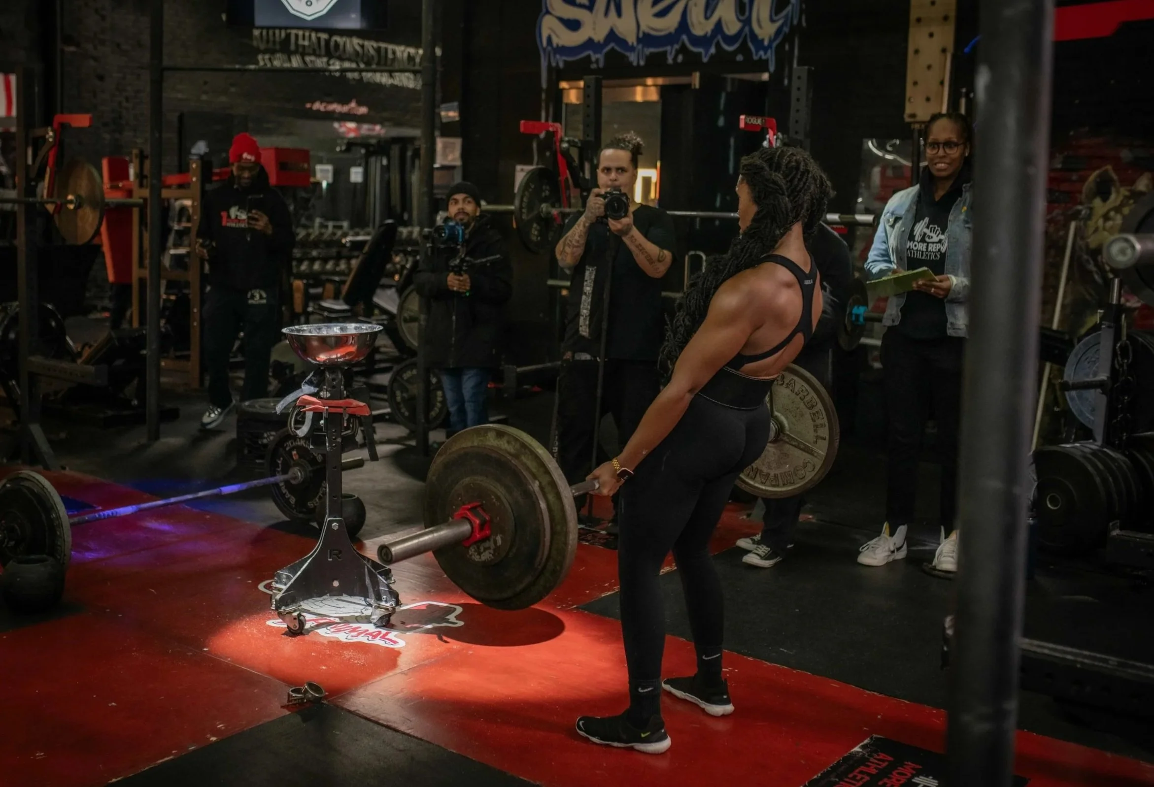 A woman in athletic wear lifting a barbell loaded with weights in a gym, surrounded by four people, some taking photos or videos and others observing. The gym has workout equipment and a sign in the background.