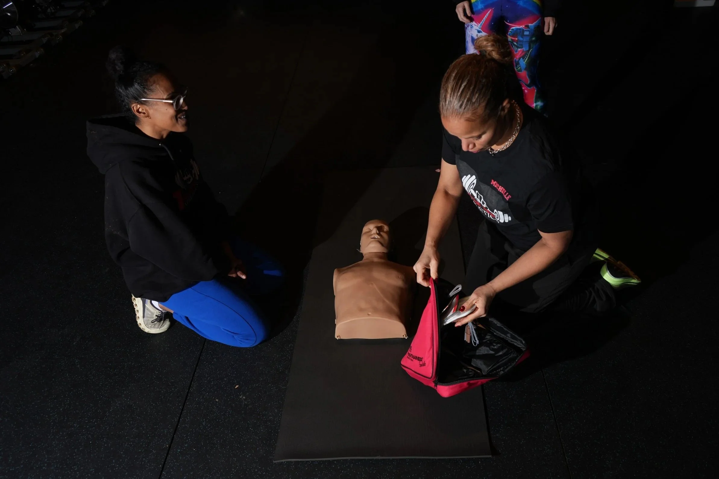 Two women practicing CPR on a training mannequin on a black mat, one kneeling and the other standing, inside a gym with black flooring.