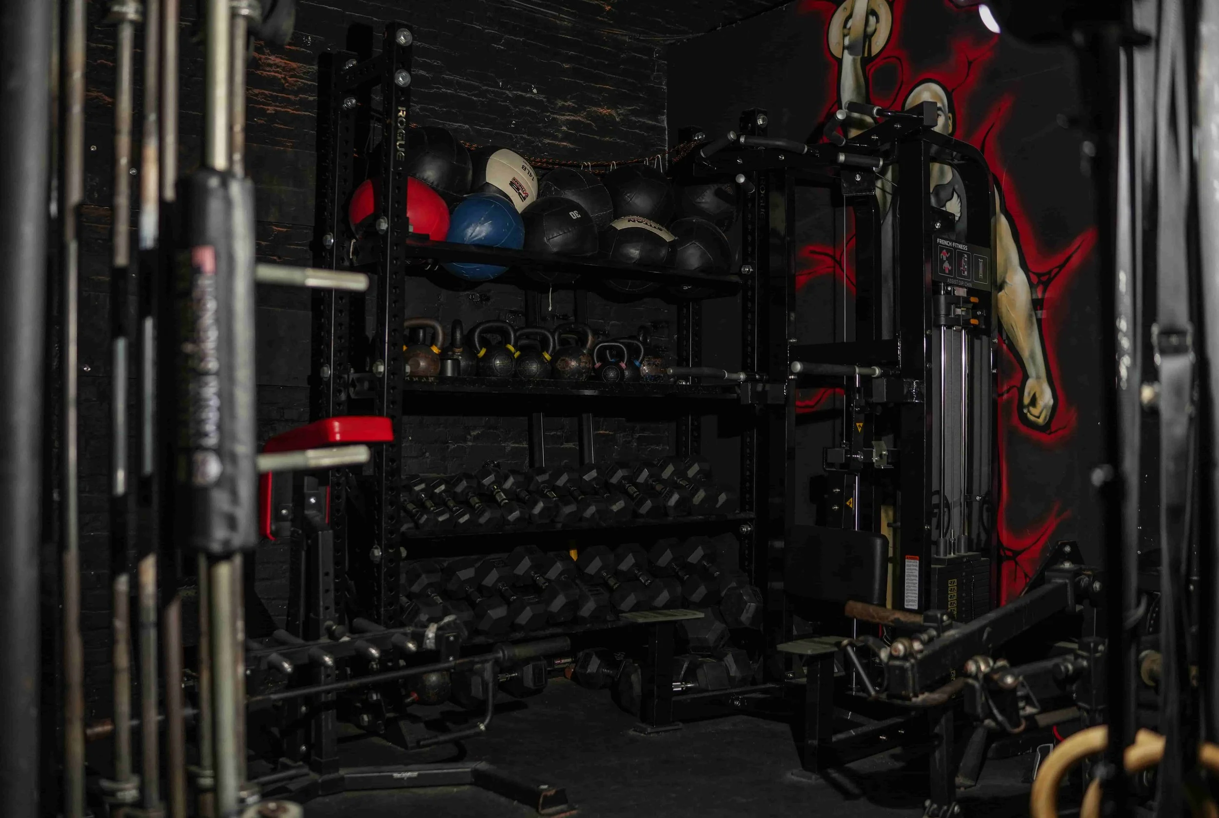 Fitness gym equipment including medicine balls, kettlebells, dumbbells, and a weight lifting machine against a black wall with a red and white graphic.