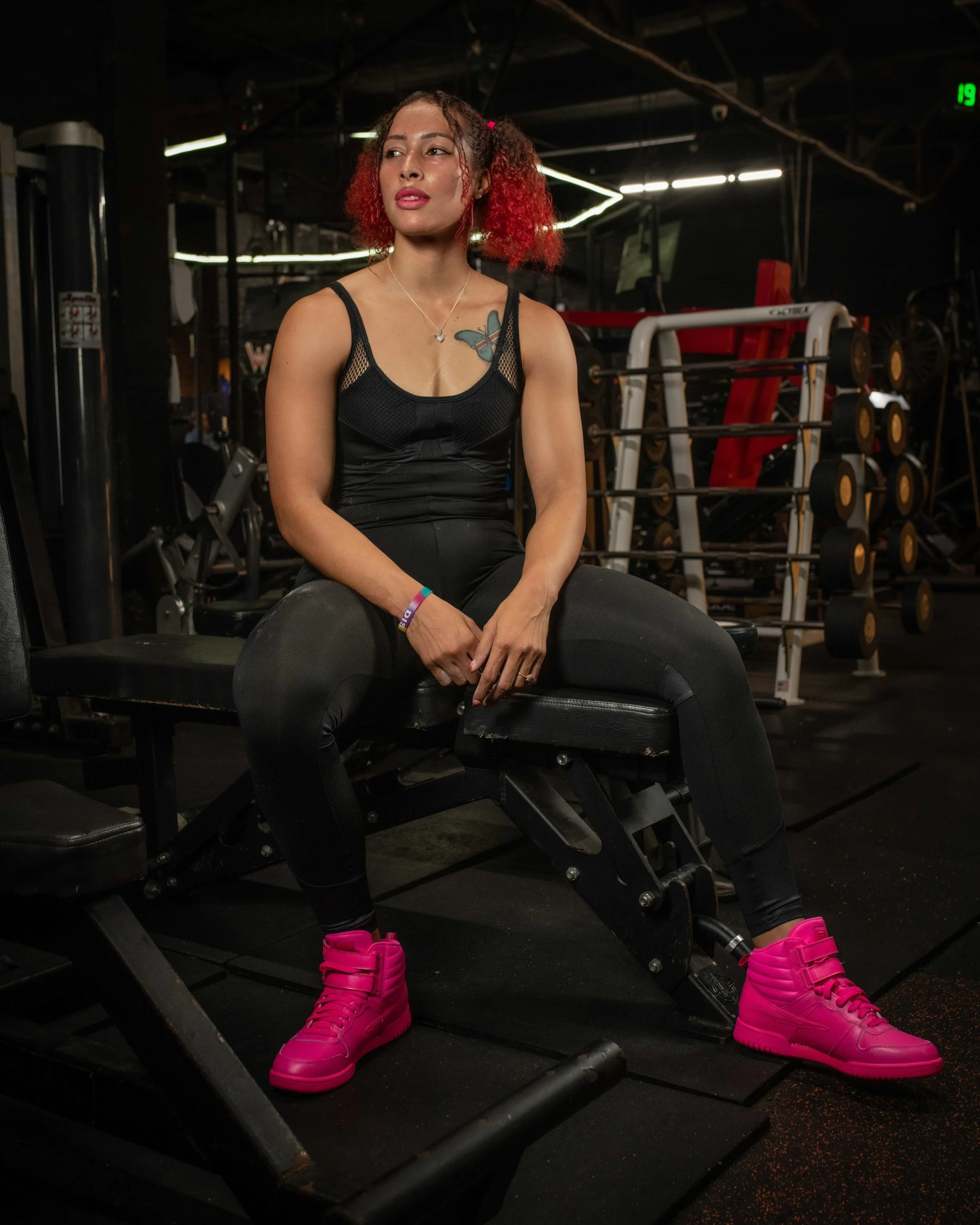 A woman with red curly hair, wearing a black workout outfit and bright pink sneakers, sitting on a bench inside a gym with weights and fitness equipment in the background.
