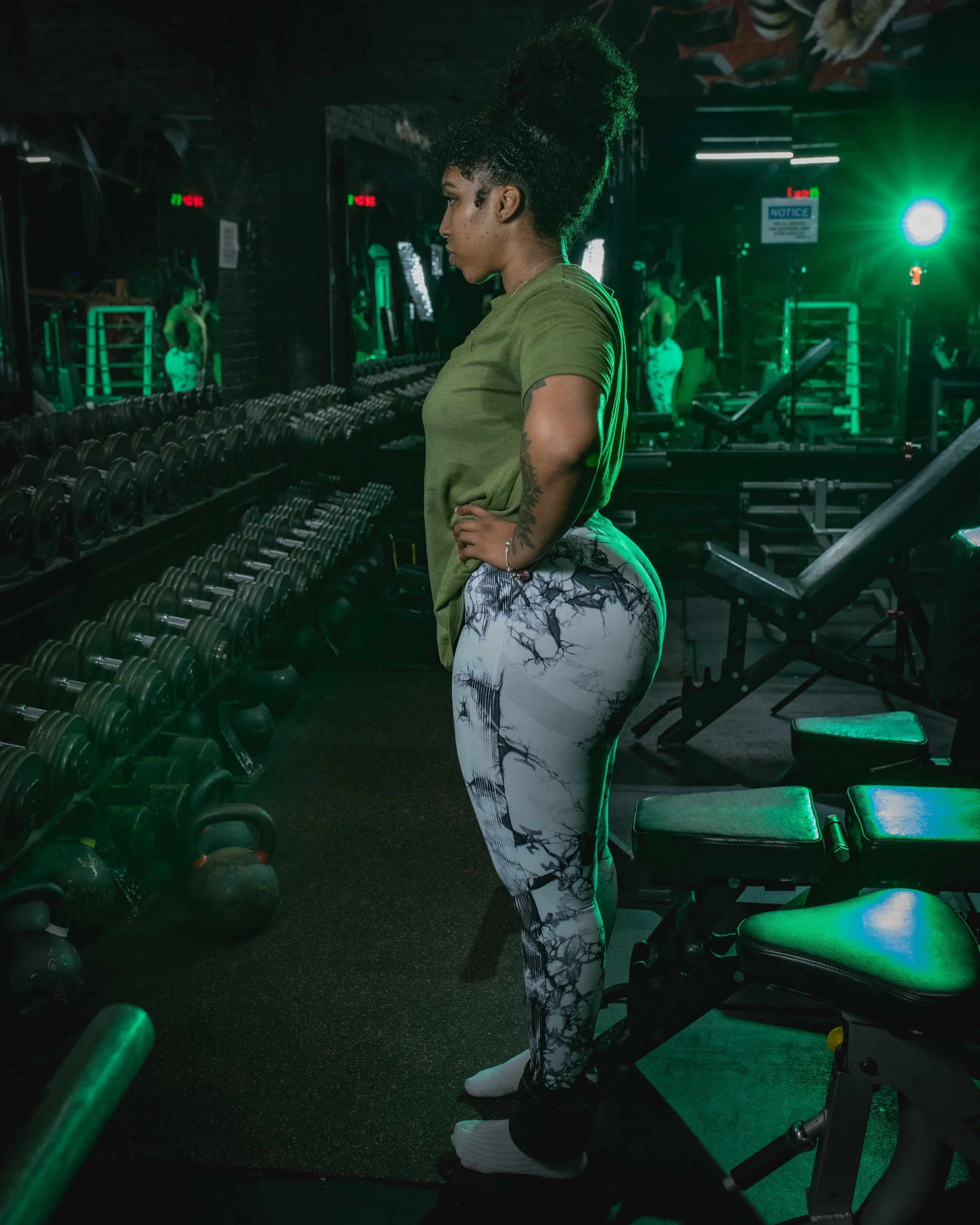 A woman in a green t-shirt and tie-dye leggings standing in a gym near a wall of dumbbells and exercise equipment, with yoga mats and kettlebells visible.