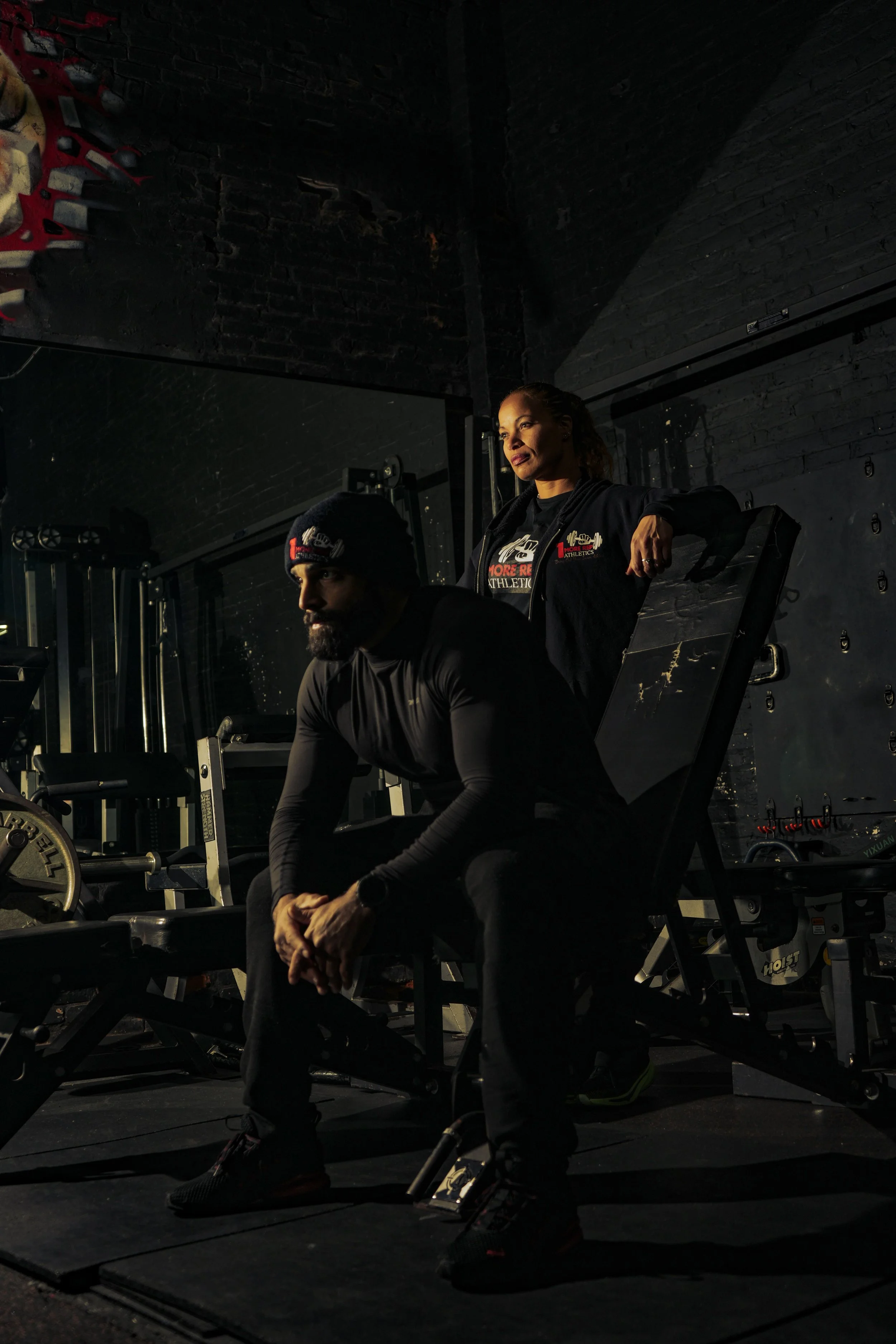 A woman leans on a gym bench while a man sits on the bench with his hands clasped, in a dark gym with black walls and equipment around them.