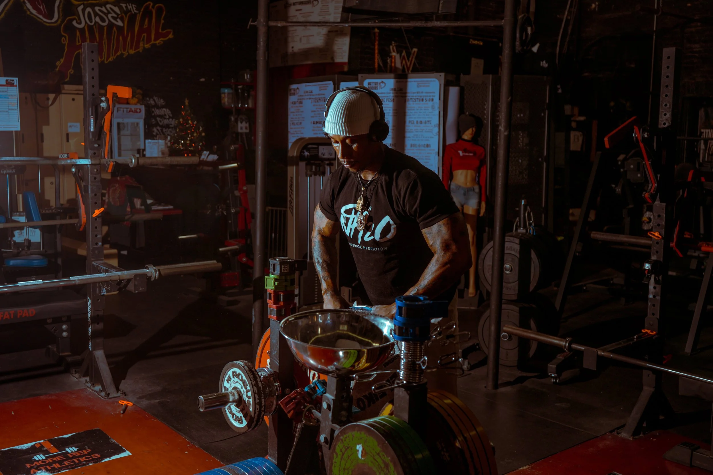 A man in a gym wearing a beanie, headphones, sunglasses, and a black t-shirt, lifting weights in a dimly lit workout space.