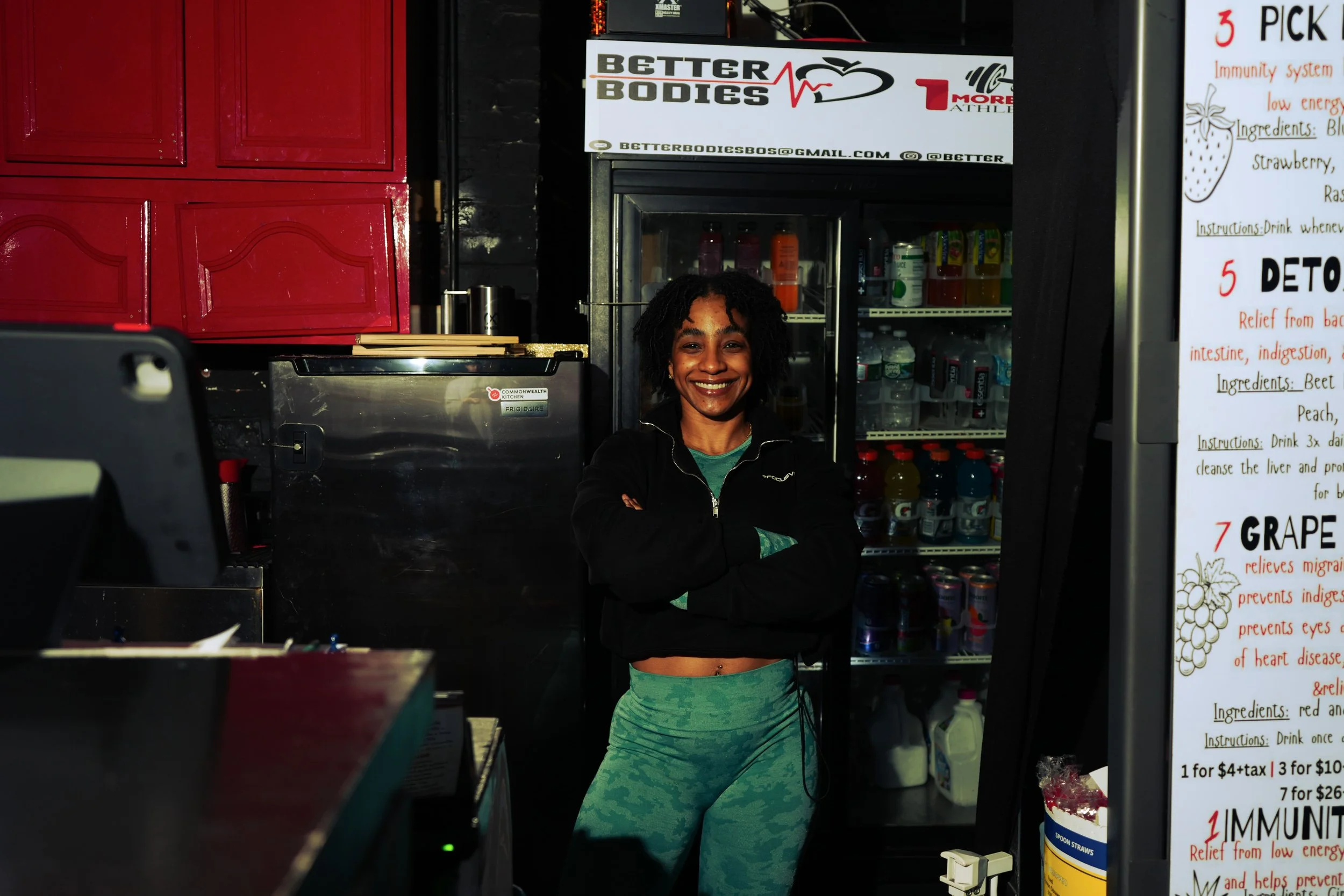 Smiling woman standing with her arms crossed in front of a refrigerator at a health food or juice bar, with a sign overhead reading 'Better Bodies'.