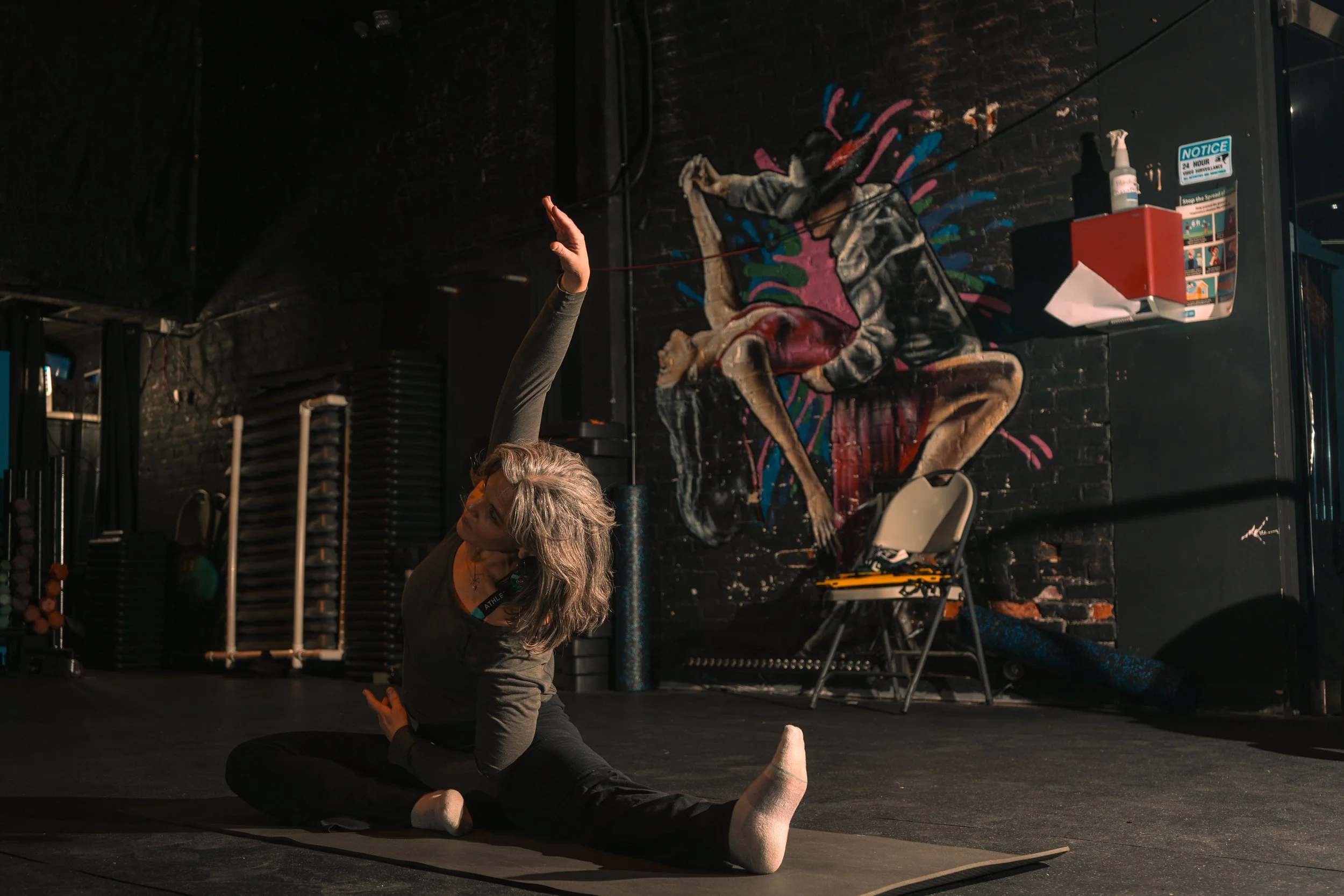 A woman practicing yoga on a mat in a dimly lit gym, with a colorful graffiti mural of a woman in a hoodie in the background.