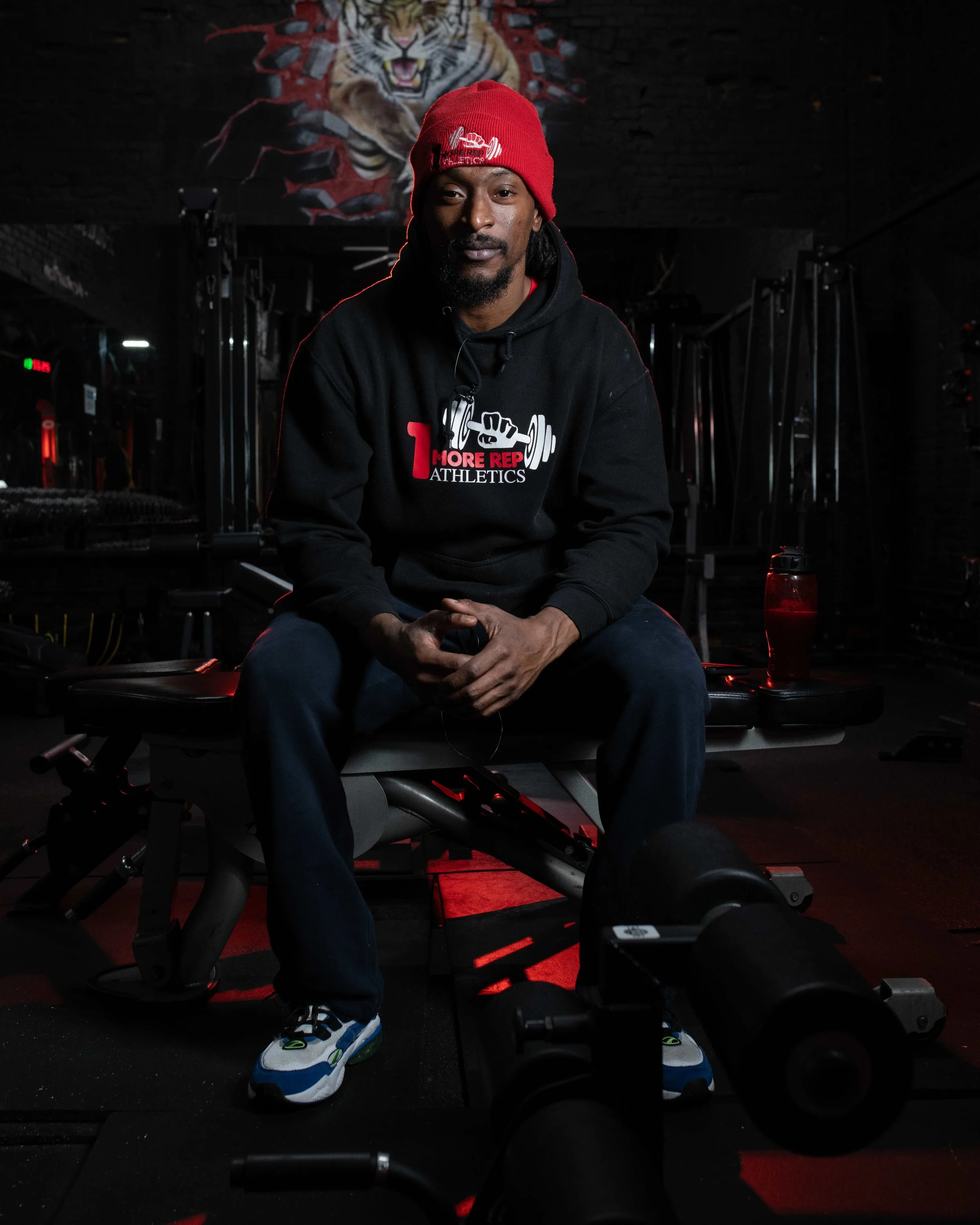 A man wearing a red beanie and black hoodie sitting on a workout bench in a gym, with gym equipment in the background and a tiger mural on the wall.