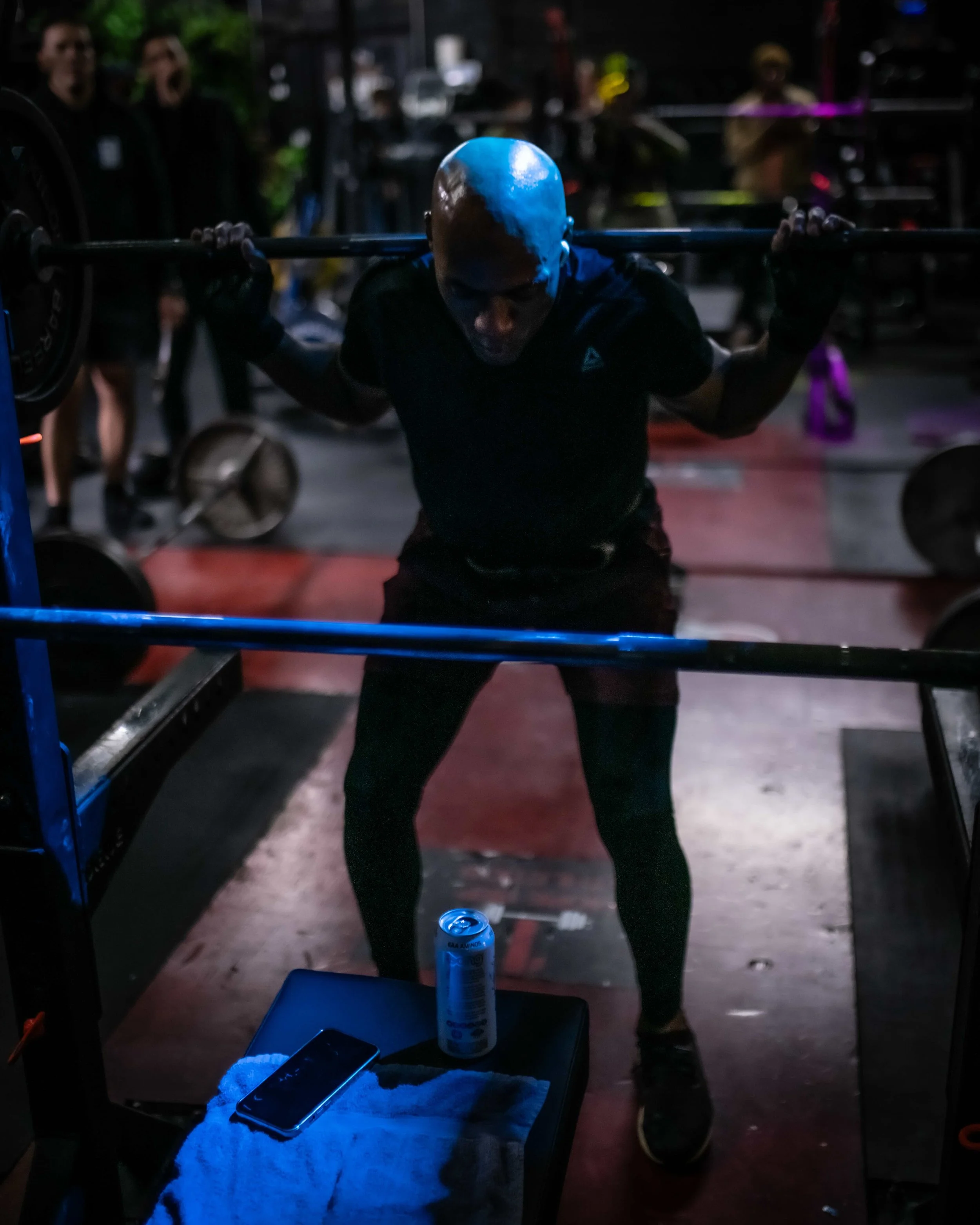 A man doing a squat with a barbell on his shoulders in a gym, with people and equipment in the background.