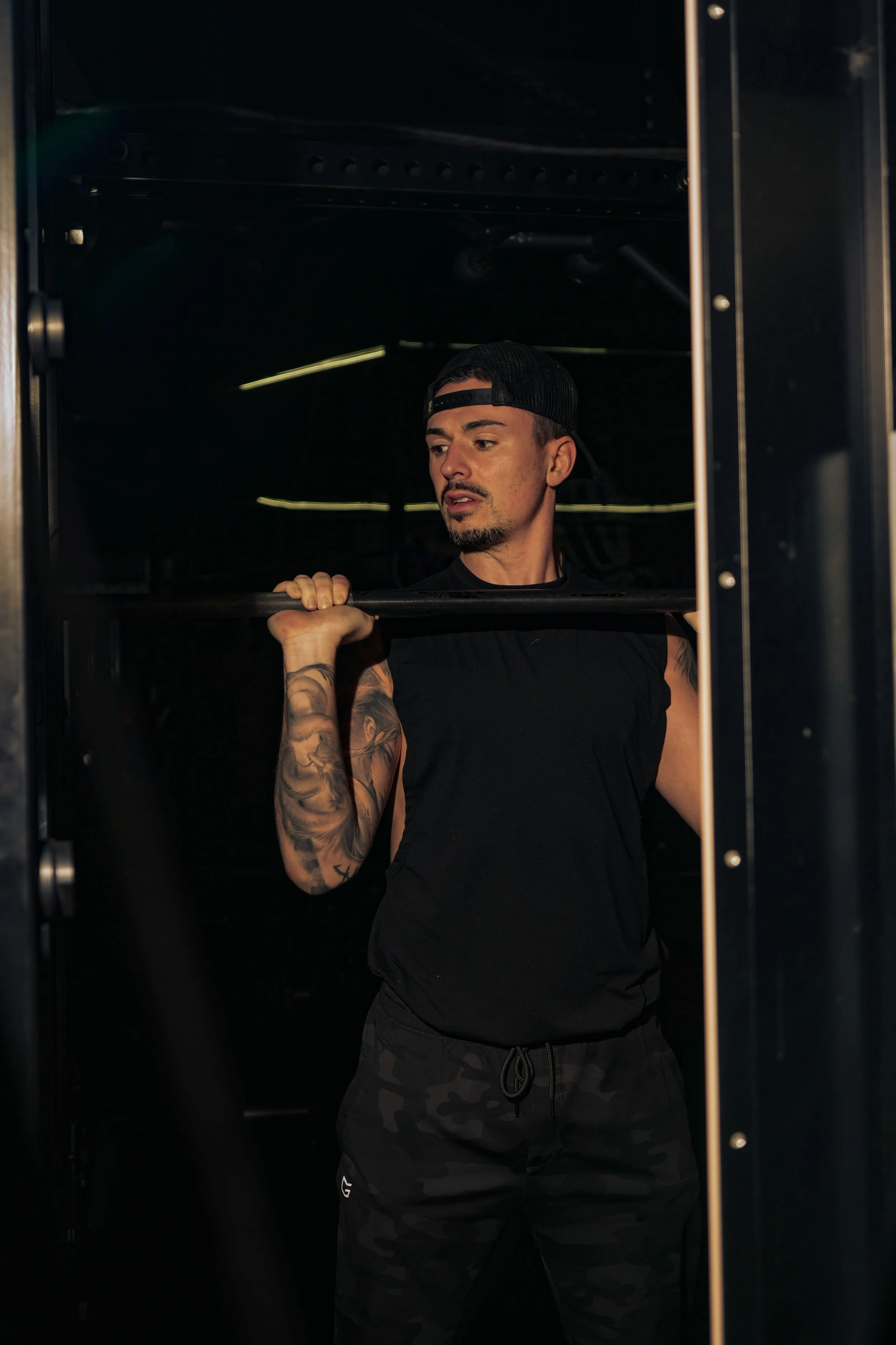 A man in a black tank top, camo workout pants, and a black backward cap is lifting a barbell in a gym. He has tattoos on his left arm and appears to be focused.