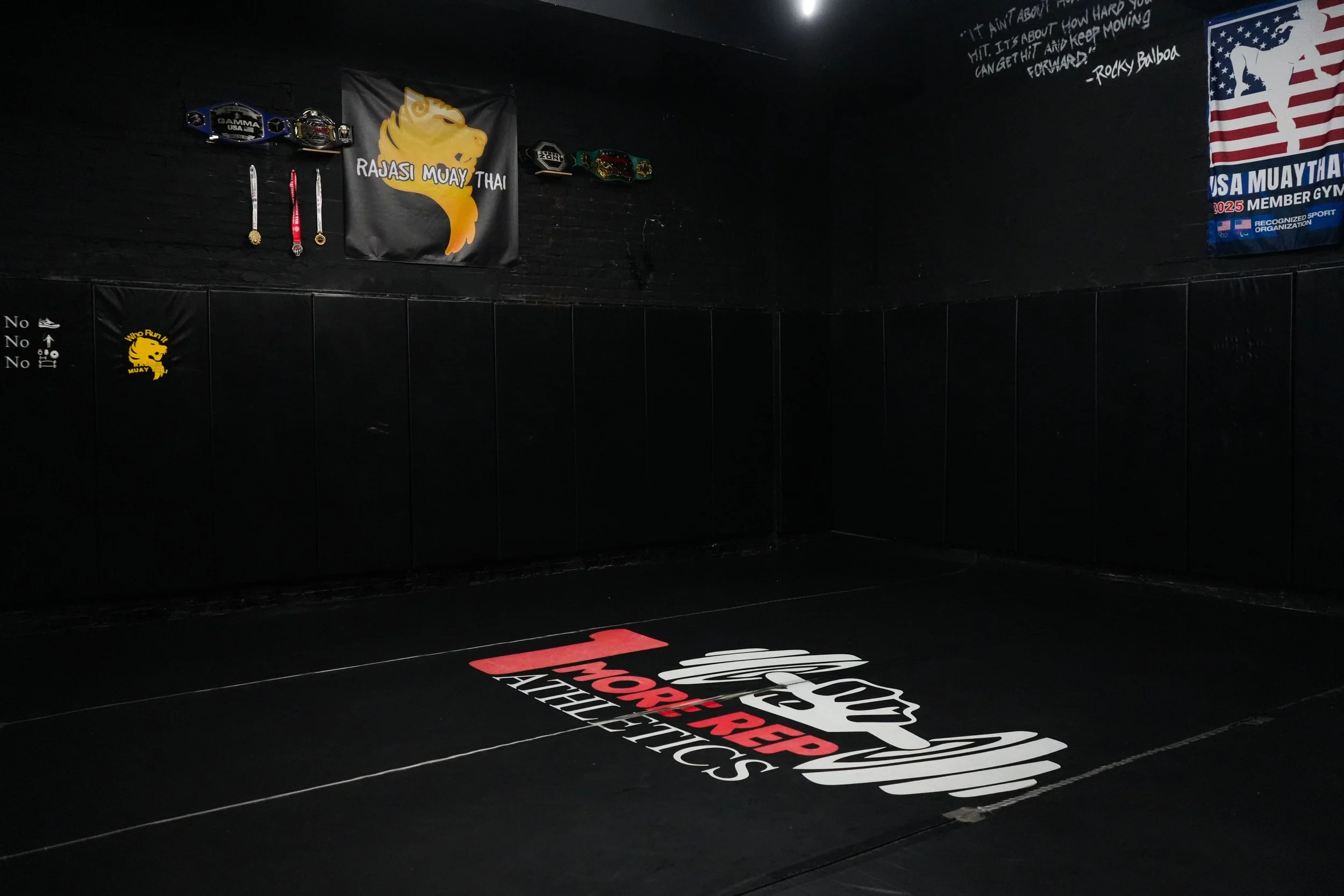 An empty MMA training room with black padded floors and walls, featuring banners with logos for Muay Thai, medals, and championship belts displayed on the wall.