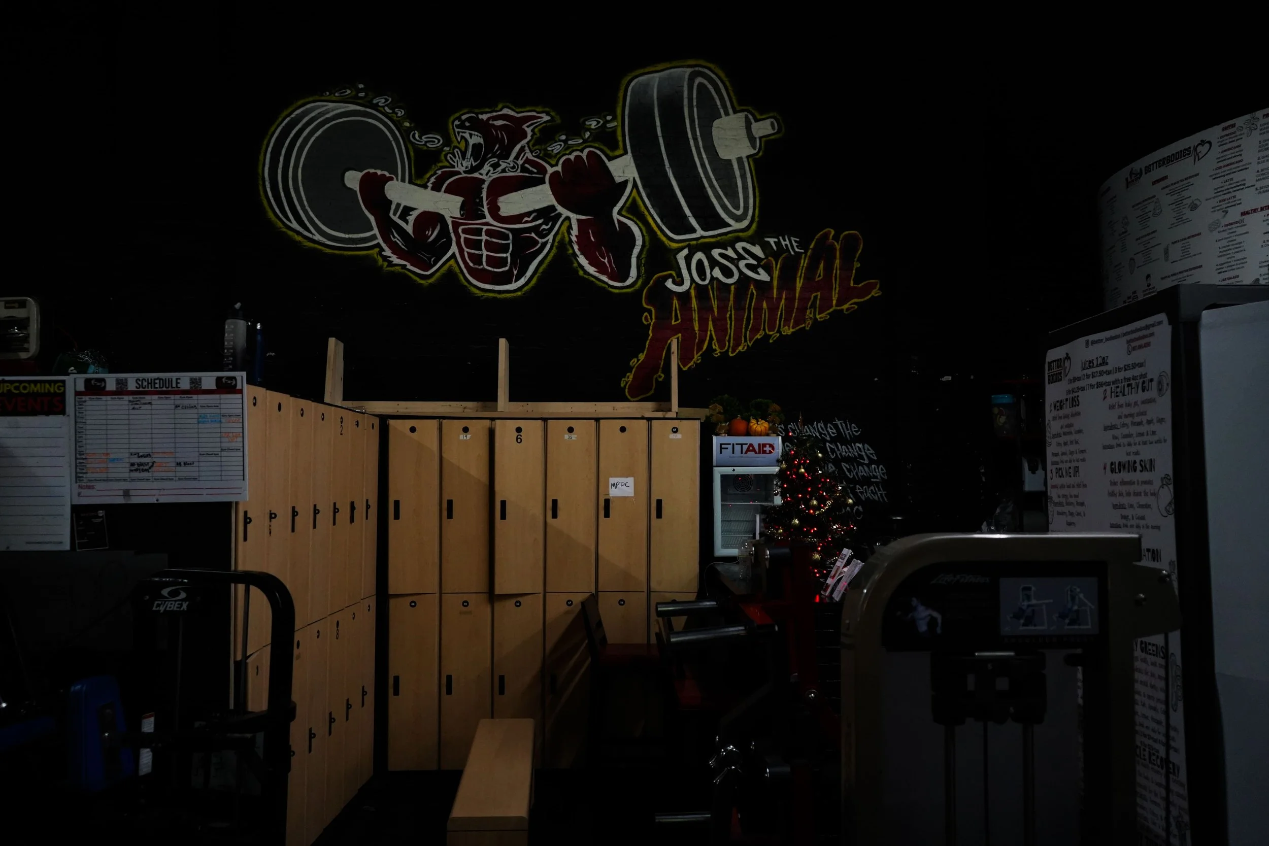 A gym with lockers, Christmas tree, gym equipment, and a wall mural of a muscular character lifting a barbell, with the words "The Joss the Animal".