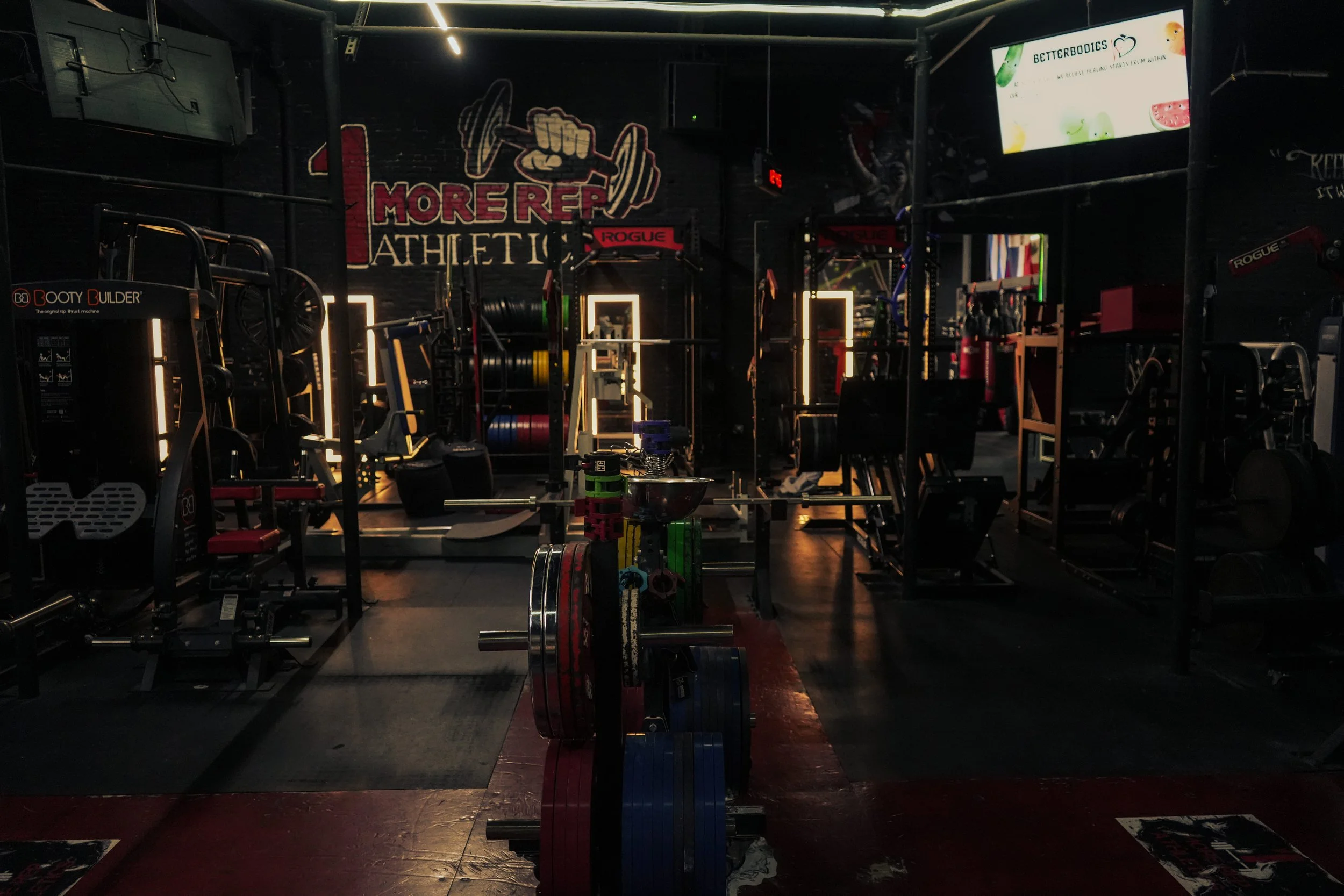 Inside a dimly lit gym with weightlifting equipment, colorful weights, and a neon sign on the back wall that reads 'More Ref Athletic'.