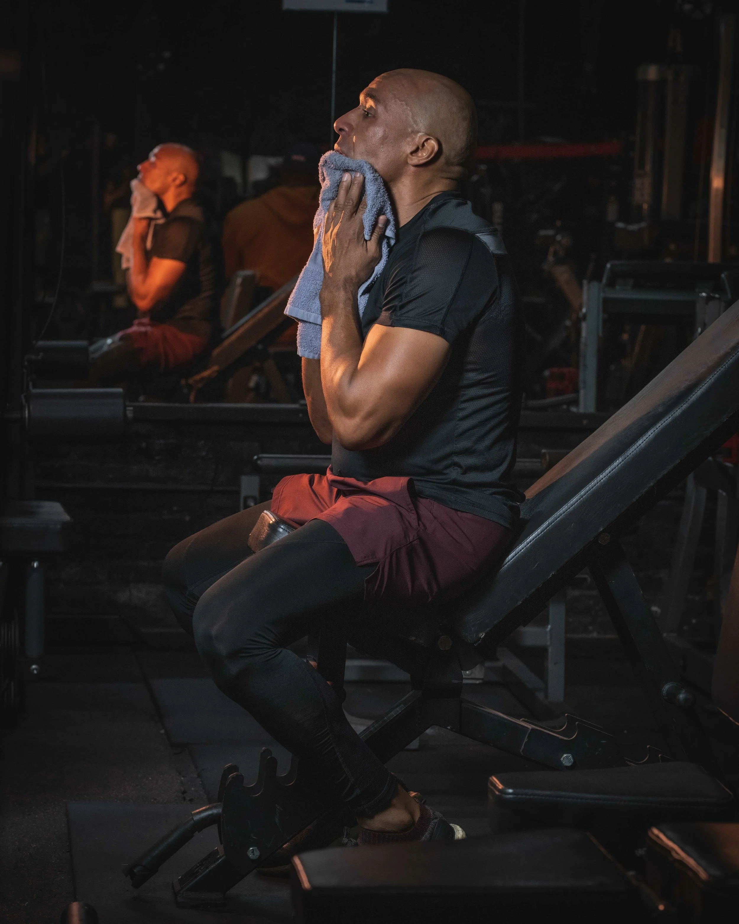 Man in gym sitting on workout bench, wiping sweat with towel, in a dark gym with equipment and mirror.