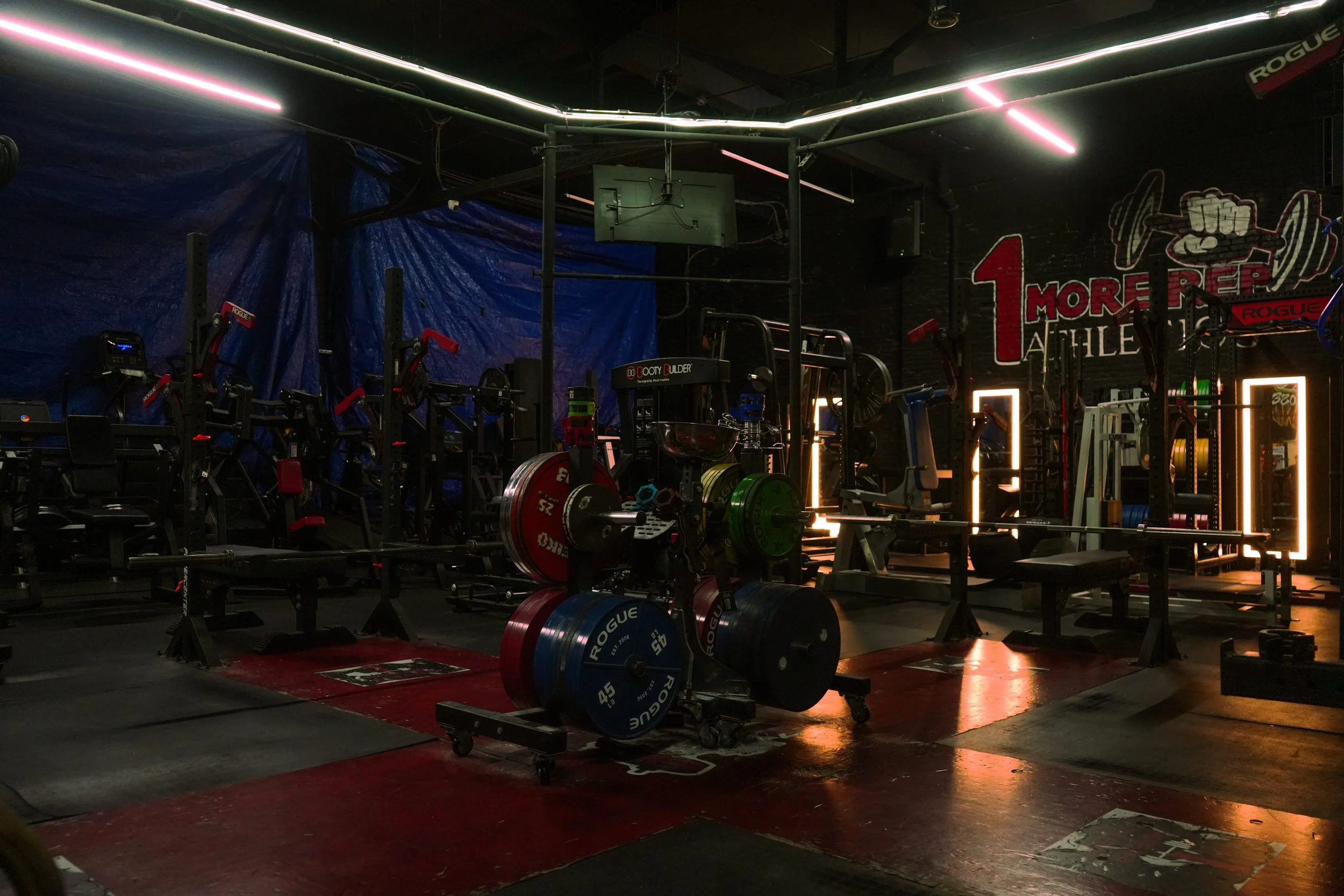 Inside a dark gym with weightlifting equipment, including barbells with colored plates, rowing machines, and squat racks, illuminated by neon pink and white lights.