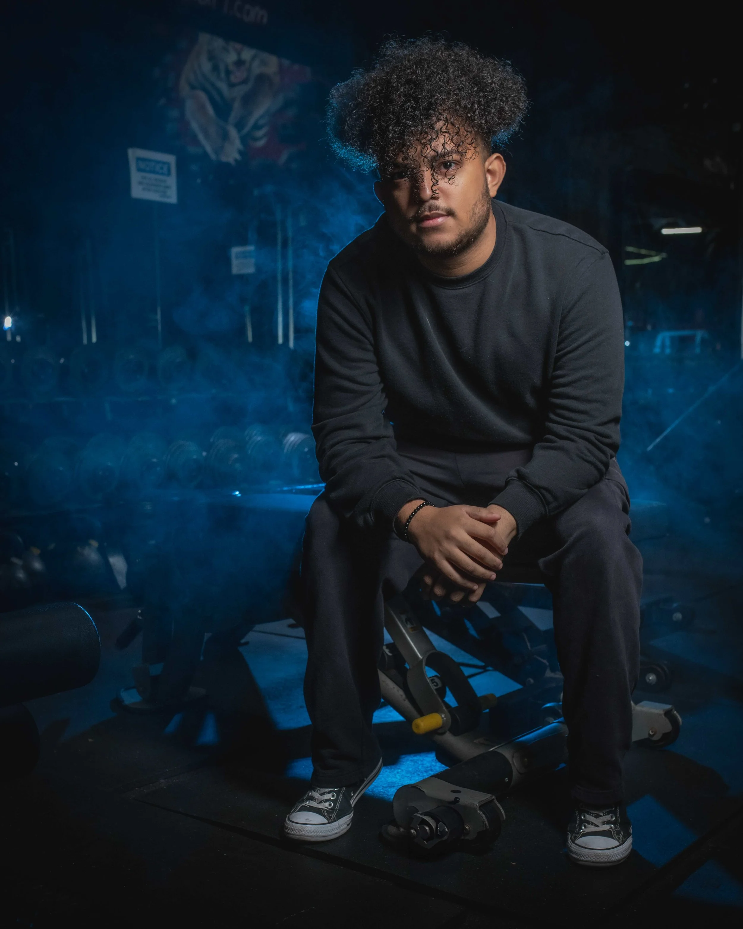 A young man with curly hair, sitting on a medicine ball in a gym, surrounded by workout equipment, with a blue smoky atmosphere.