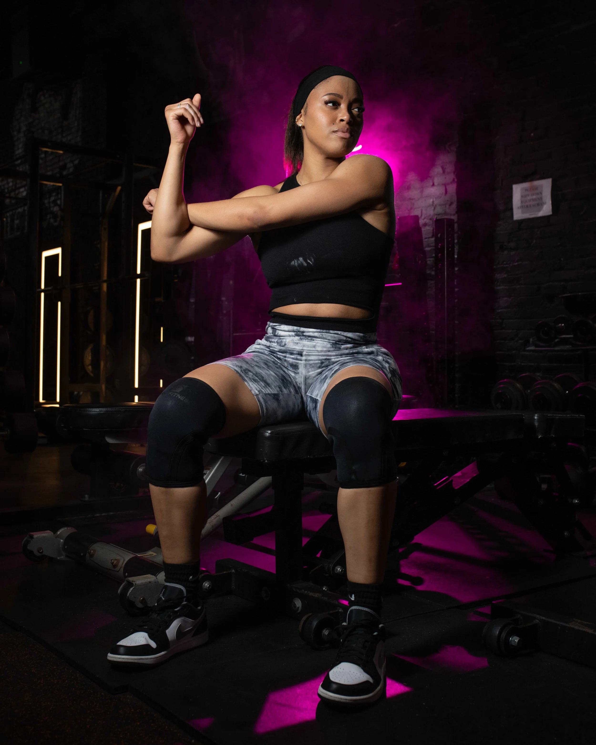 A young woman in workout attire sits on a bench at the gym, flexing her arm muscles. She is wearing a black crop top, patterned gray shorts, knee pads, and sneakers. The background features gym equipment and purple lighting.