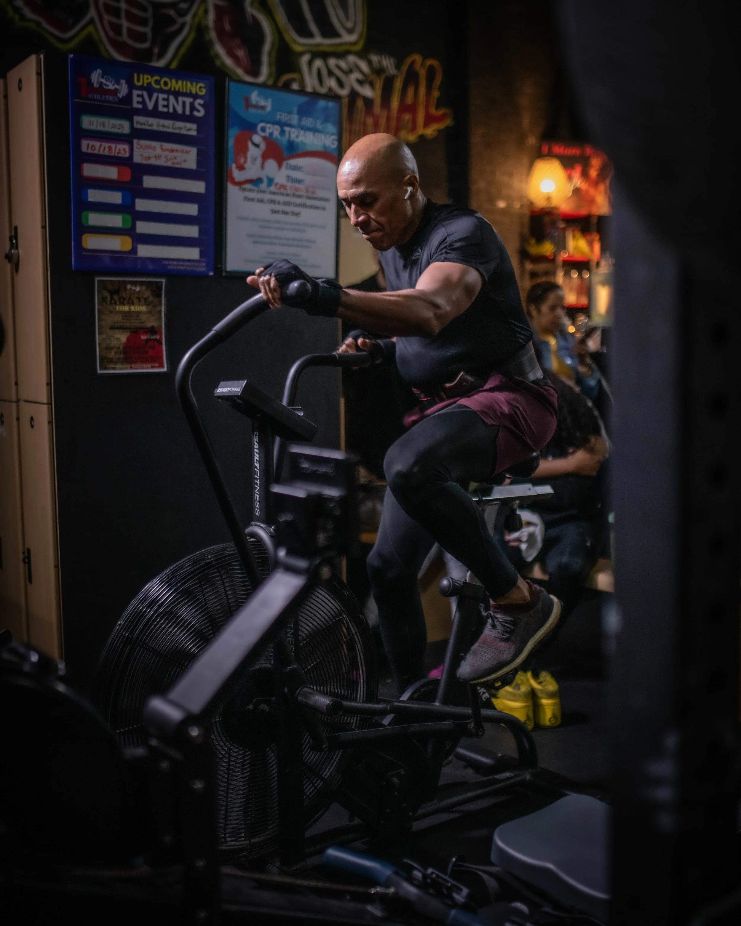 Man exercising on an air bike in a gym, with posters and other people in the background.