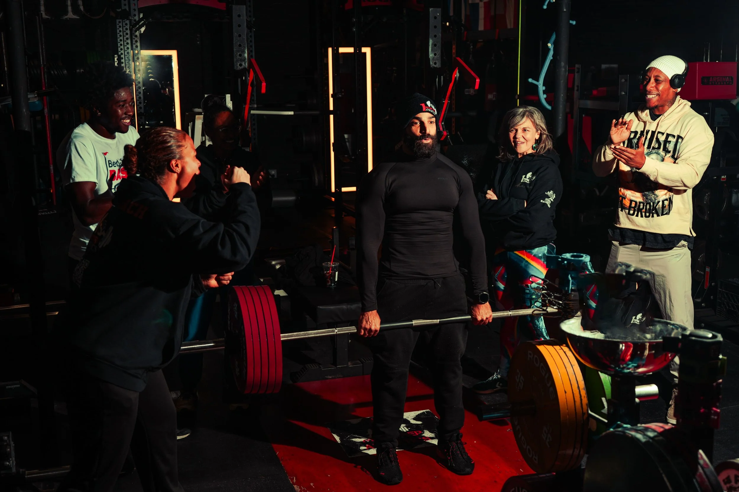 Group of people in a gym, with one person lifting weights and others clapping and smiling around him.