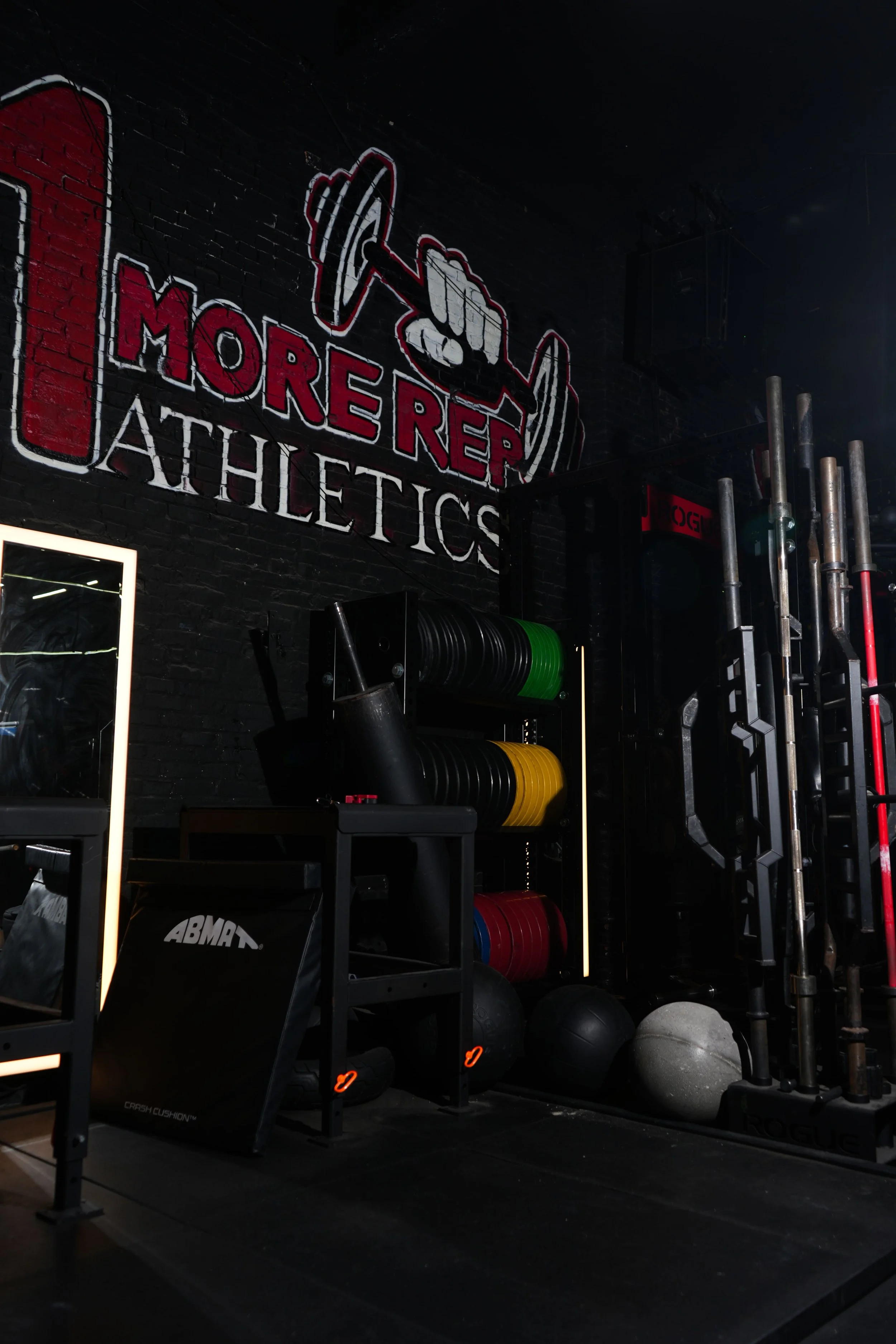 Interior of a gym with fitness equipment, a wall sign reading 'More Reps Athletics', and various weights and medicine balls.