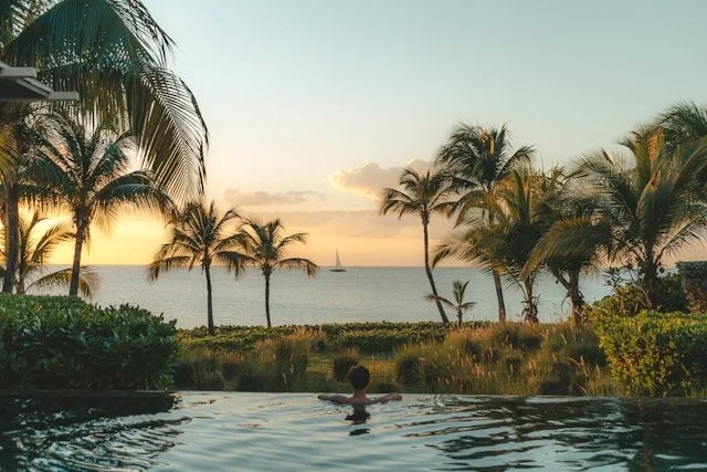 Person relaxing in a swimming pool overlooking a tropical beach with palm trees and a sunset sky.