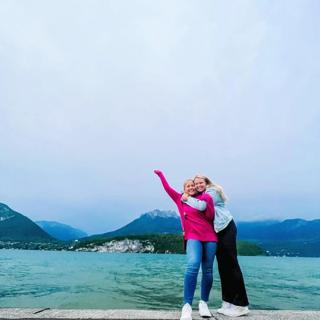 Two women hugging by a lake with mountains in the background, one woman raising her arm, during daytime with cloudy sky.