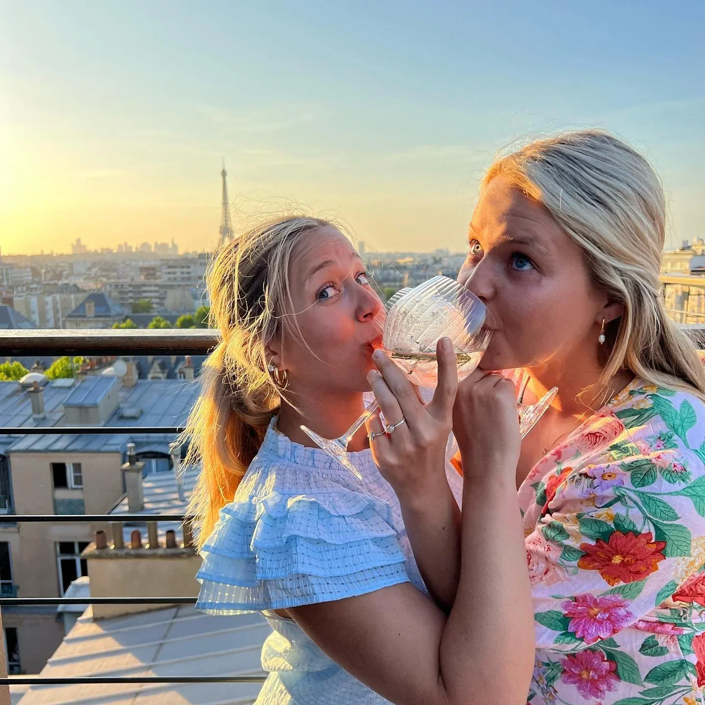 Two women with blonde hair enjoying drinks on a rooftop terrace with a cityscape and the Eiffel Tower in the background during sunset.