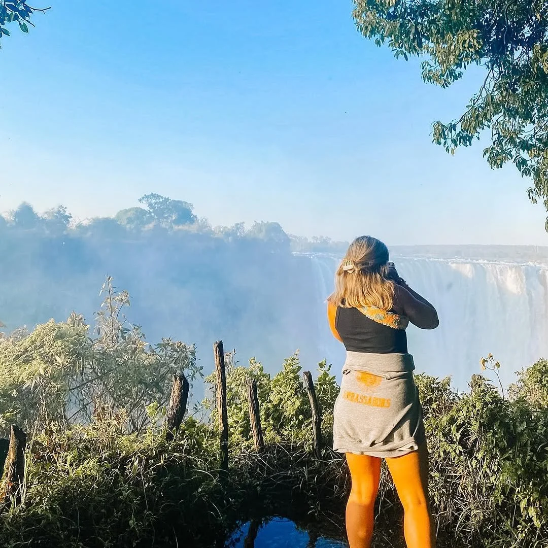 A woman with shoulder-length hair taking a photo of Niagara Falls with a camera. She is standing on a dirt path with greenery and a wooden fence in front of her. The sky is clear and blue.