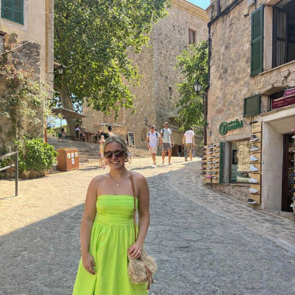 A woman wearing a bright yellow strapless dress and sunglasses standing on a cobblestone street in a historic European town. There are stone buildings, green trees, and several people walking in the background.
