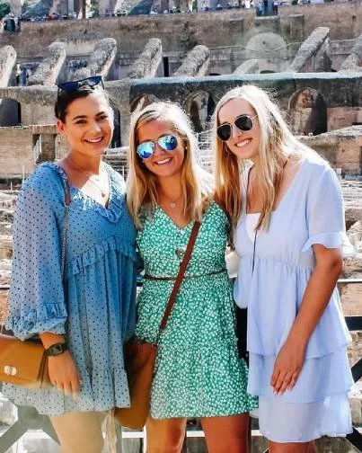 Three young women smiling outdoors near historical stone ruins, with bright attire and sunglasses.