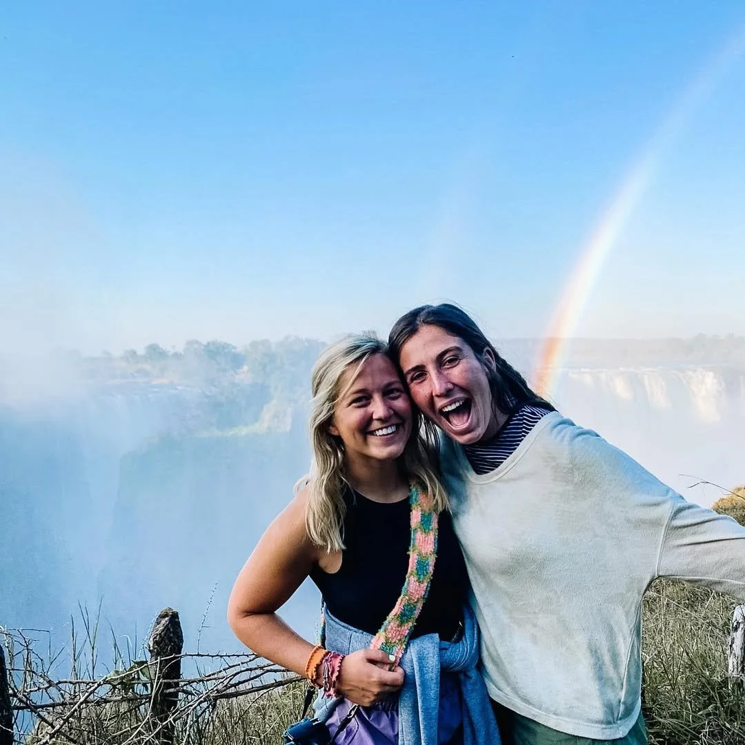Two women smiling and posing in front of Niagara Falls with a rainbow in the background.