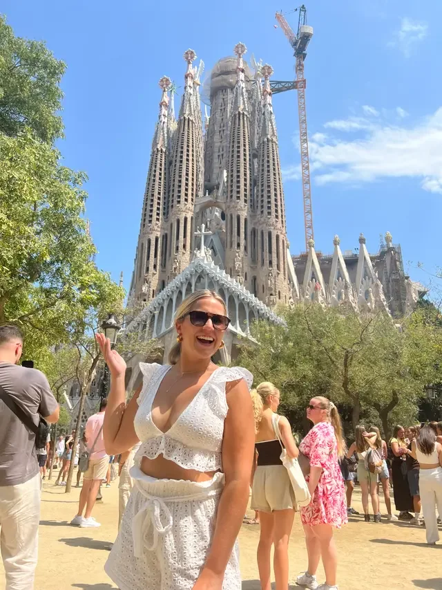 A woman wearing sunglasses and a white lace outfit smiling and pointing at the Sagrada Familia in Barcelona, Spain, during daytime with a clear blue sky and other people around.