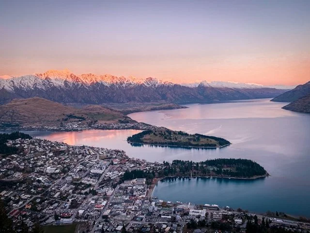 A cityscape with a lake and snow-capped mountains at sunset.
