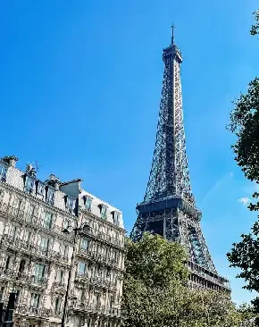 The Eiffel Tower in Paris viewed from the ground with surrounding buildings and trees under a clear blue sky.