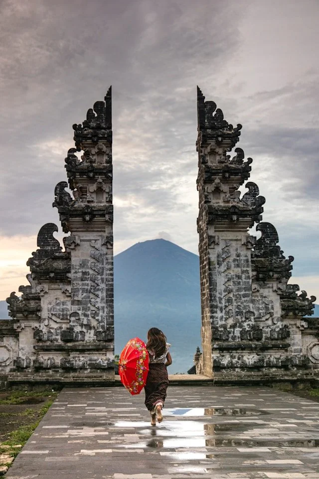 A woman with a red umbrella walking through an ornate stone gate toward a volcano in the background under a cloudy sky.