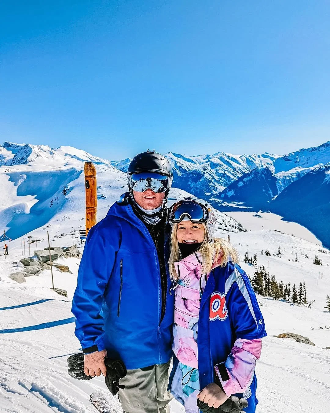 A man and woman in ski gear standing on a snowy mountain with a scenic backdrop of snow-covered mountains and a clear blue sky.