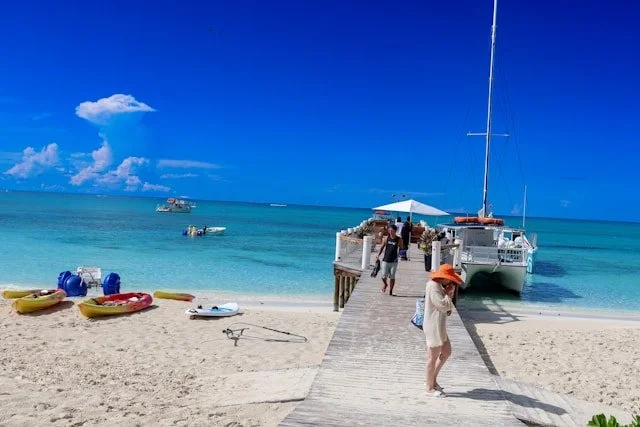 A beach scene with a wooden dock extending into clear blue water, with a sailboat moored at the dock, kayaks and small boats on the sand, and people walking and relaxing under a white umbrella.