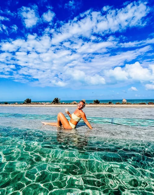 Woman in a yellow bikini sitting on the edge of a swimming pool with a view of the ocean and blue sky filled with clouds.