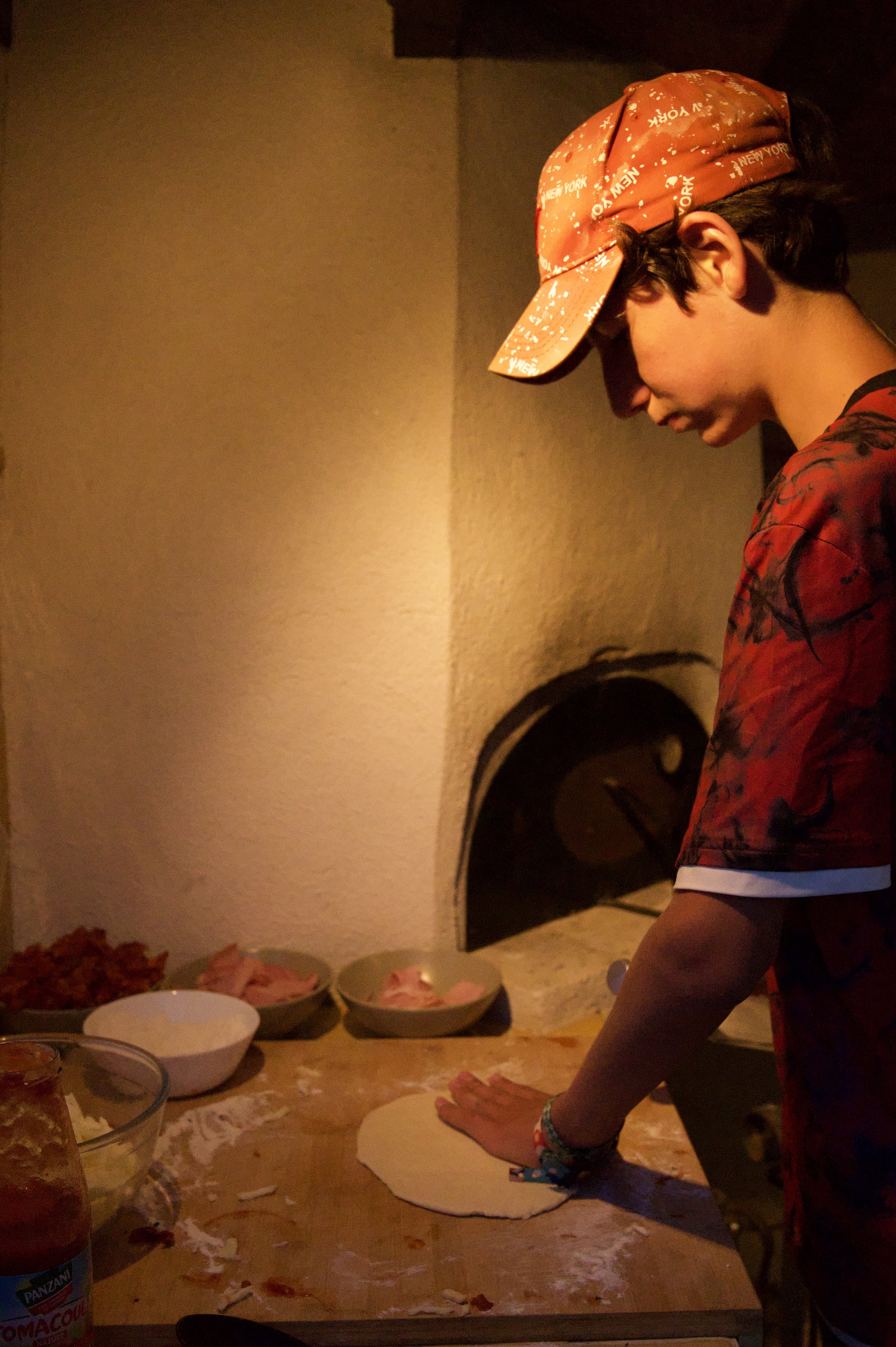 Une personne portant un bandana orange et un t-shirt à motifs rouges prépare des ingrédients pour une pizza dans une cuisine chaleureuse, avec plusieurs bols contenant de la nourriture sur une table en bois.