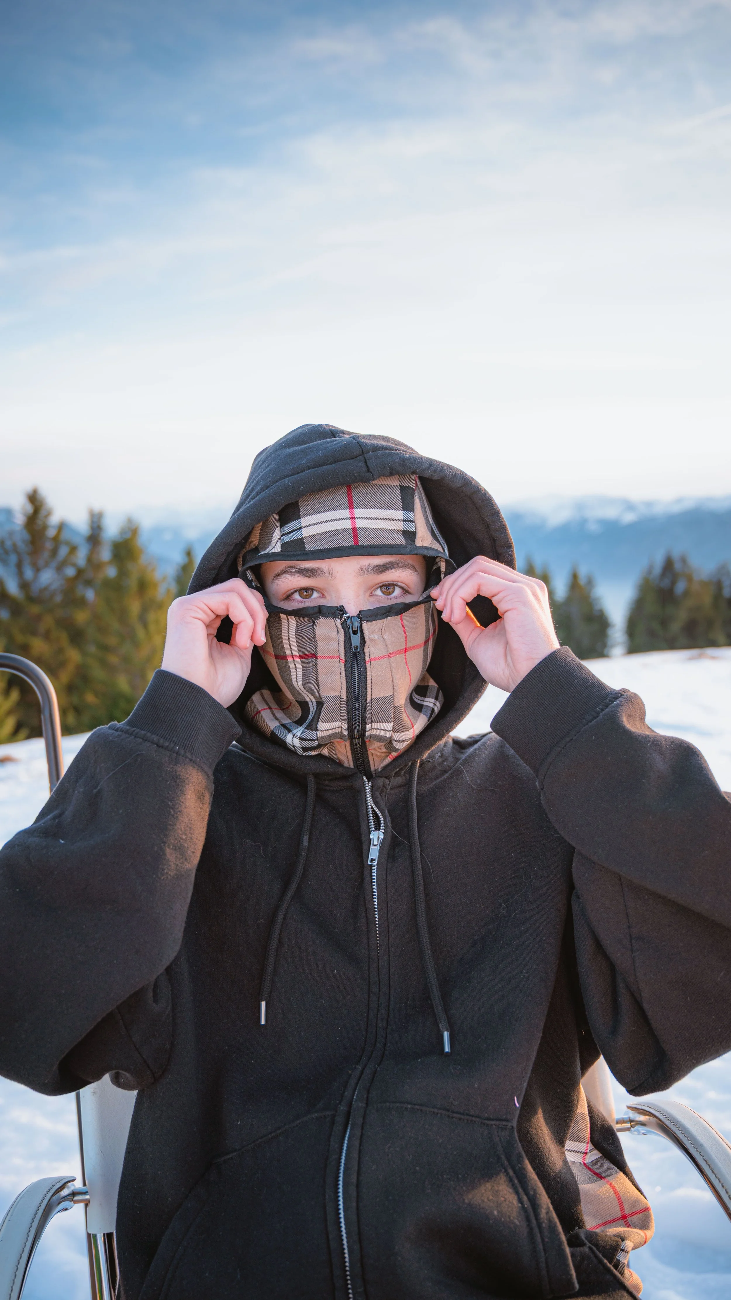 Jeune homme assis en fauteuil sur neige, portant un casque, une capuche et un masque, avec des montagnes en arrière-plan.