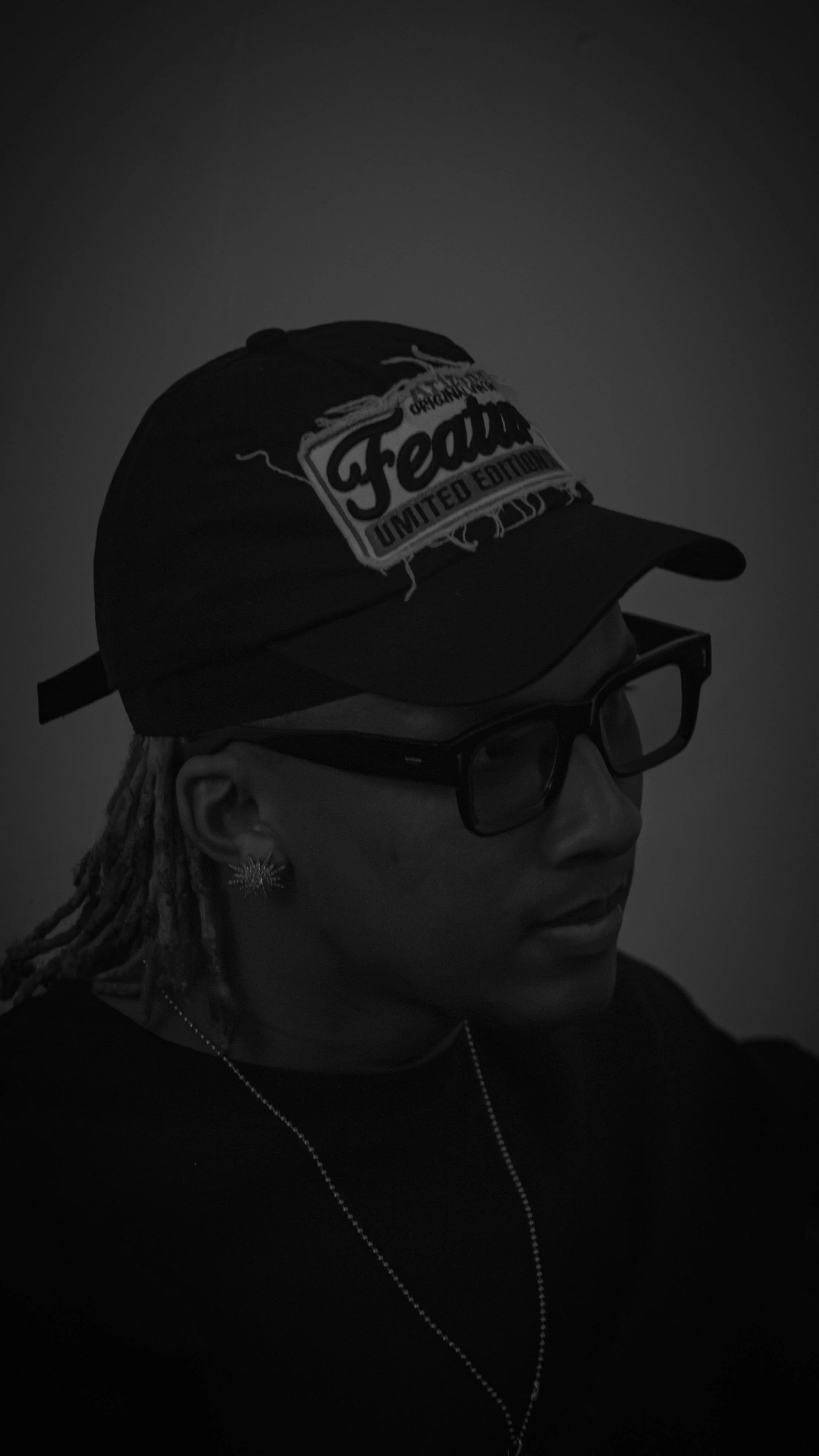 Une personne portant une casquette noire avec un logo blanc et noir, des lunettes noires épaisses, une boucle d'oreille en forme d'étoile, des dreadlocks, et un collier, vue en profil dans une image en noir et blanc.
