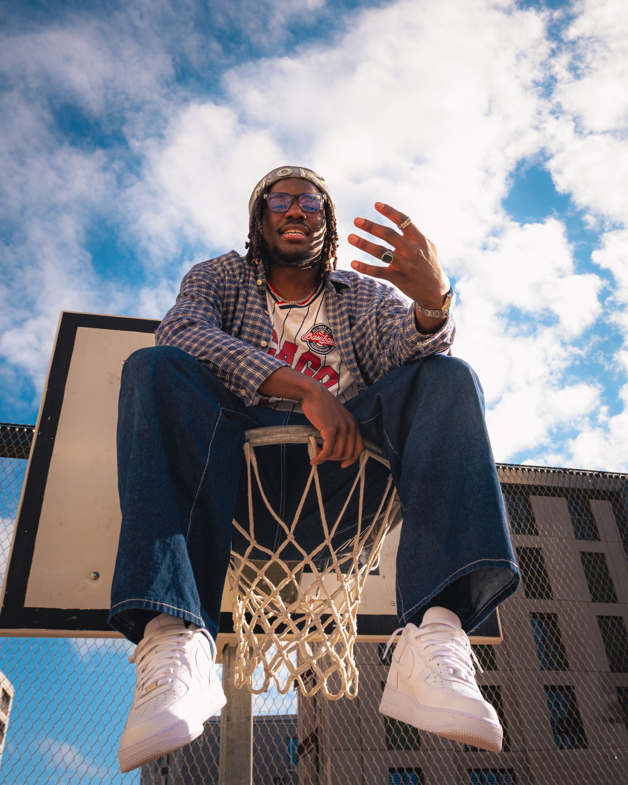 Person assise sur un panier de basketball vue de dessous, portant des baskets blanches, un jean et un haut de sport. Le ciel bleu avec des nuages blancs en arrière-plan.