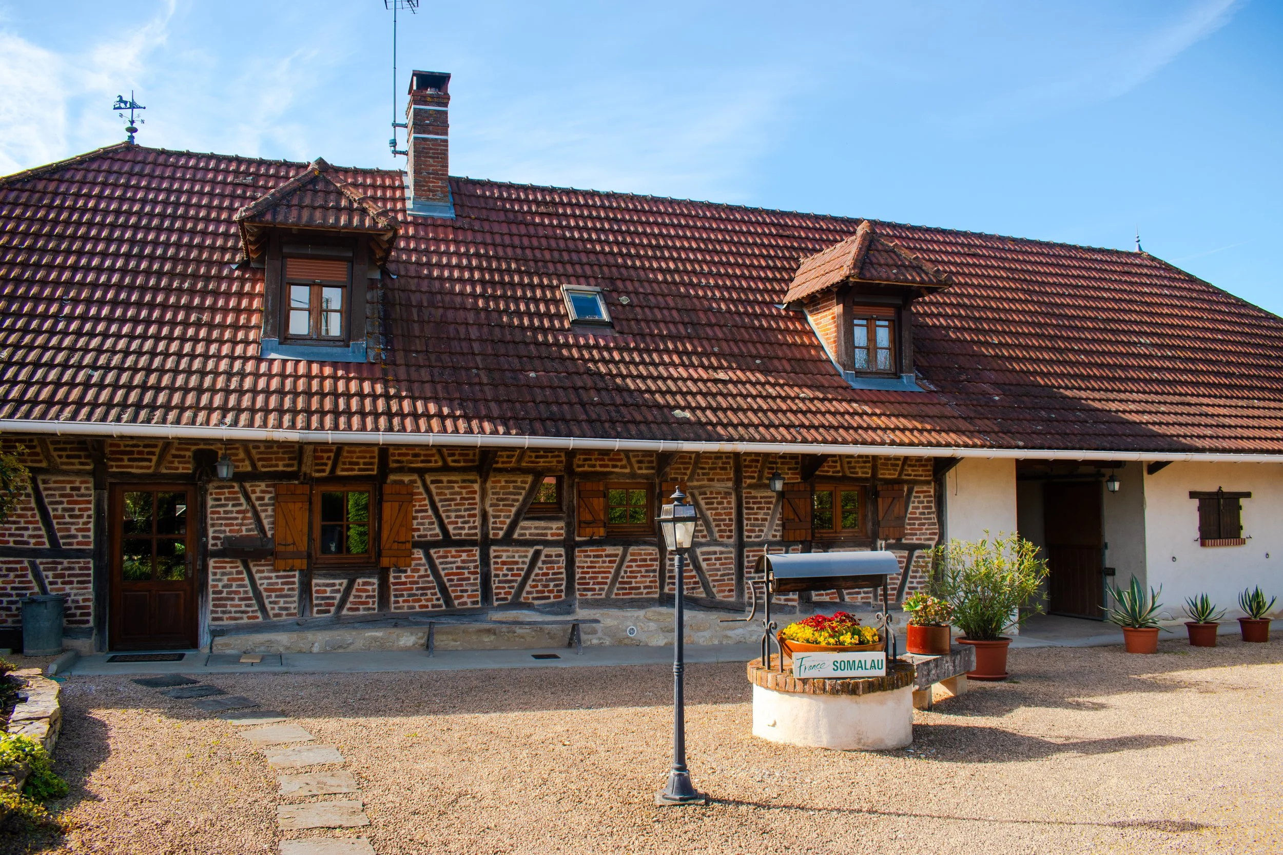 Une maison en pierre avec un toit en tuiles rouges, fenêtres à volets en bois, et un jardin devant. Il y a aussi une boîte aux lettres et un panneau indiquant 'France SOMALAU'.