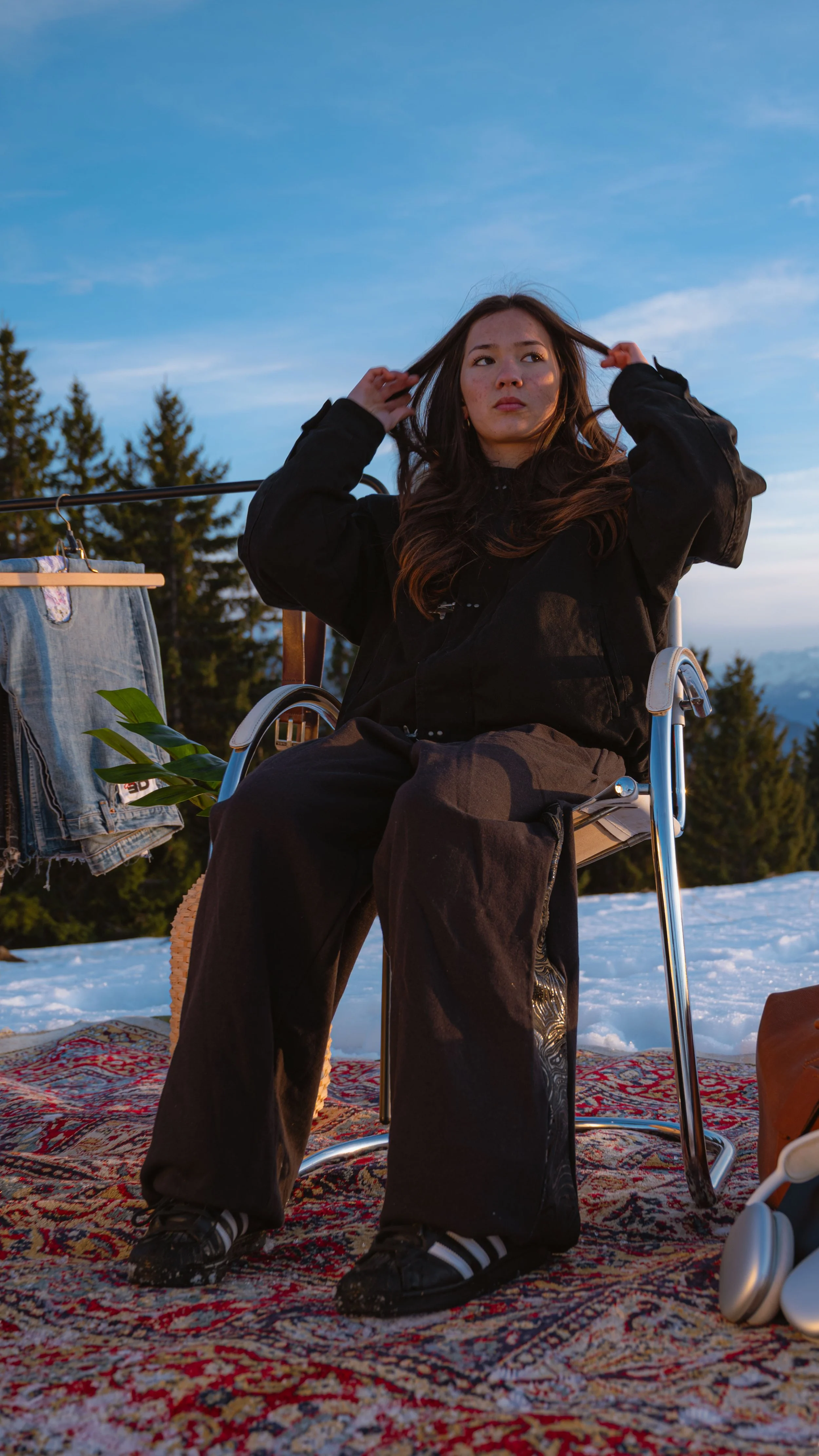 Jeune femme en fauteuil roulant, portant un manteau noir, s'assoit sur une couverture tissée dans un environnement extérieur enneigé avec des arbres verts en arrière-plan, en train de jouer avec ses cheveux, sous un ciel bleu.