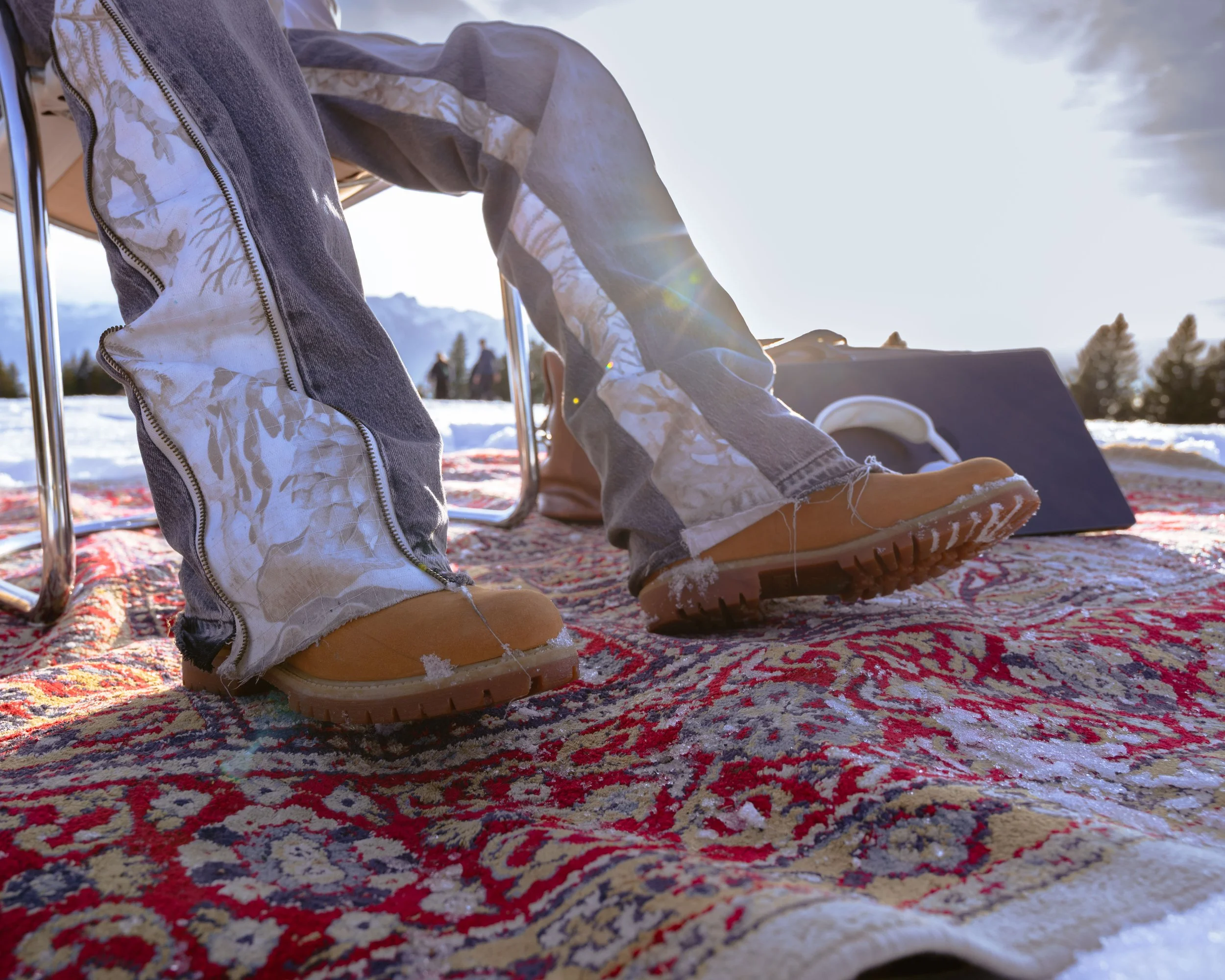 Personnes assises sur un tapis de style persan, chaussées de bottes d'hiver en bois, dans un paysage enneigé avec des montagnes et des arbres au loin.