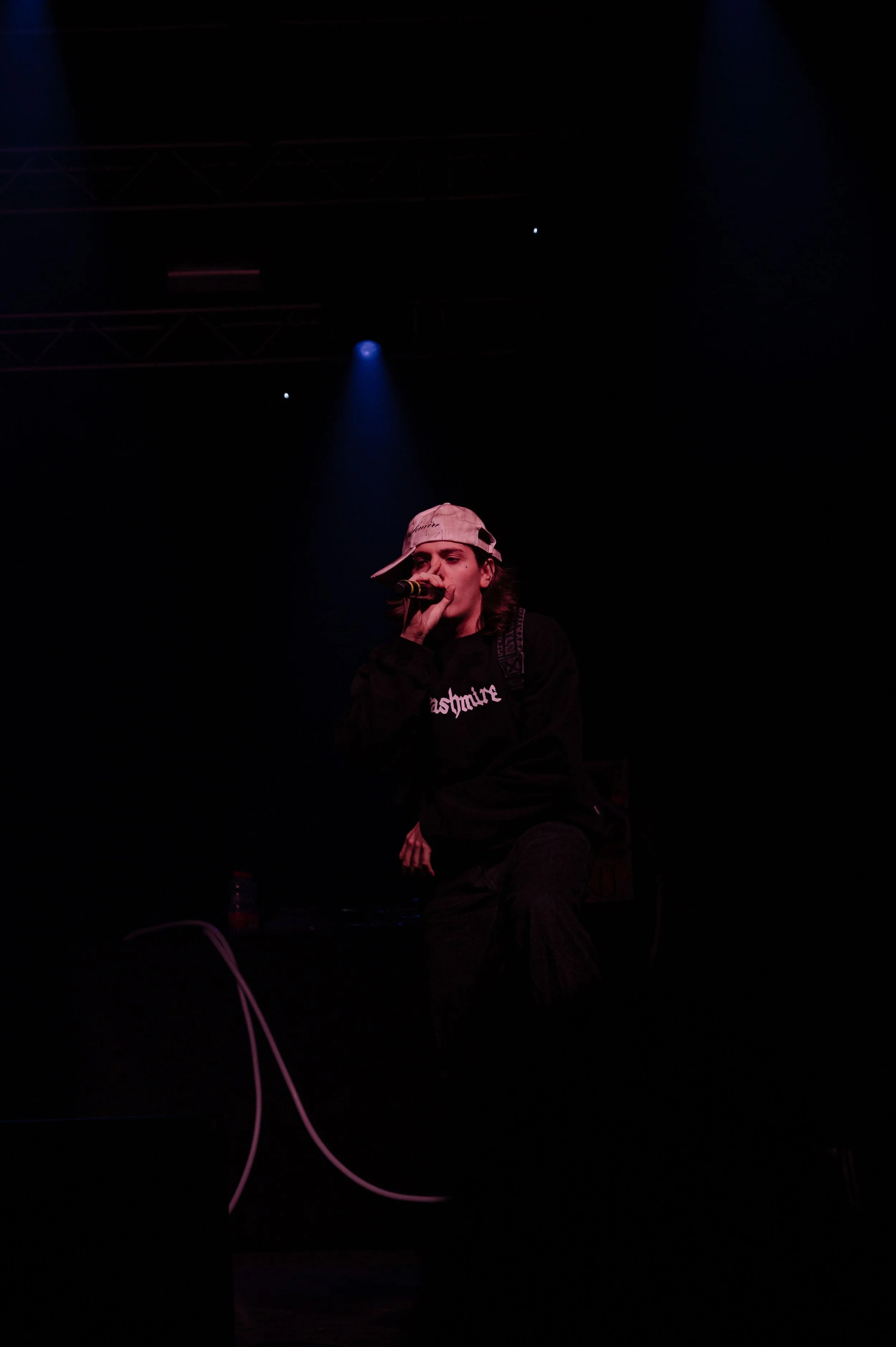 Un jeune homme chante dans un microphone sur scène, portant une casquette décalée, dans un environnement sombre avec des lumières de scène.
