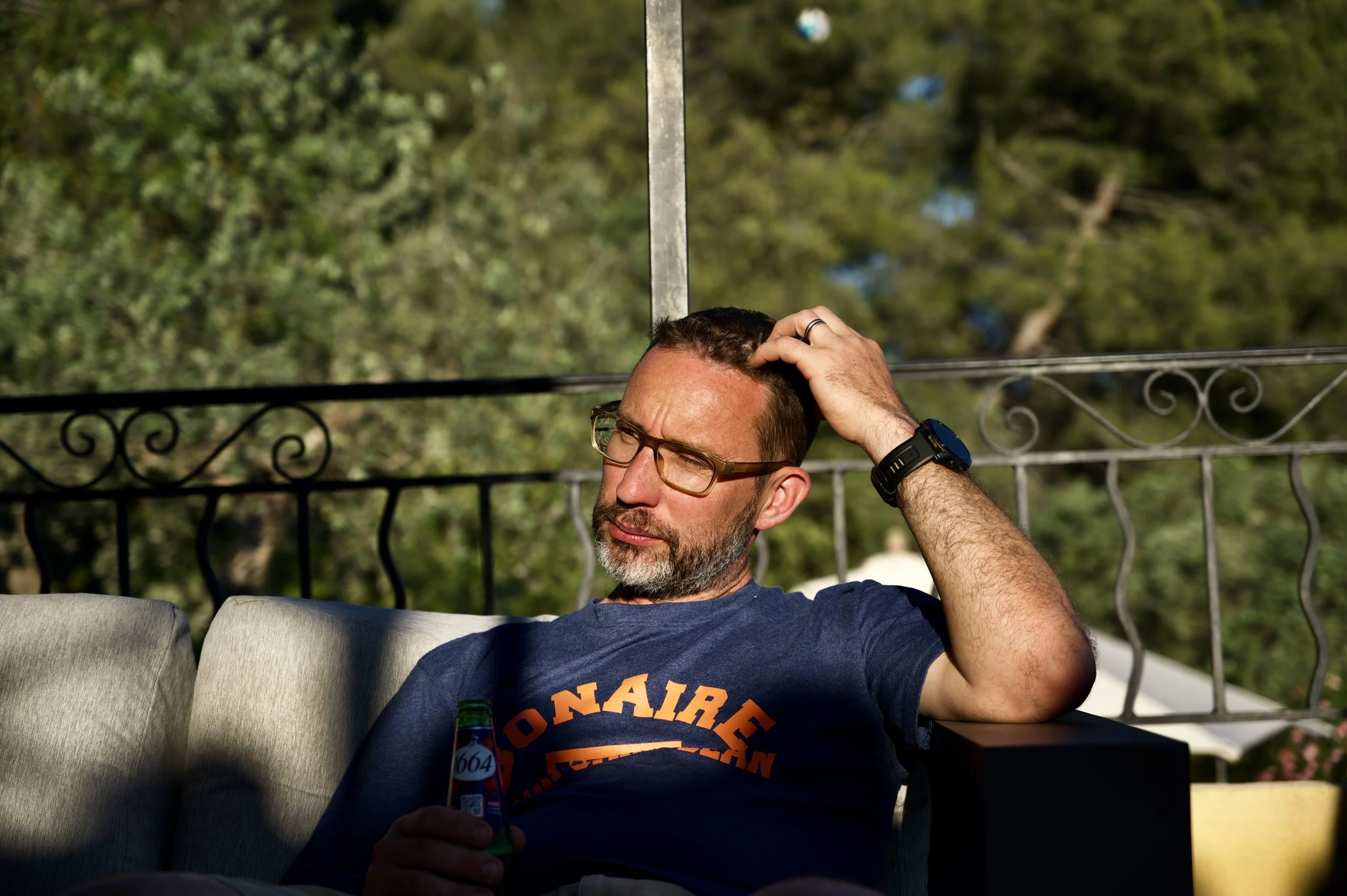 Homme assis sur un canapé, portant des lunettes, un t-shirt et un bracelet noir, tenant une bouteille de bière, avec un fond de décoration de balcon ou terrasse en fer forgé, et des arbres en arrière-plan
