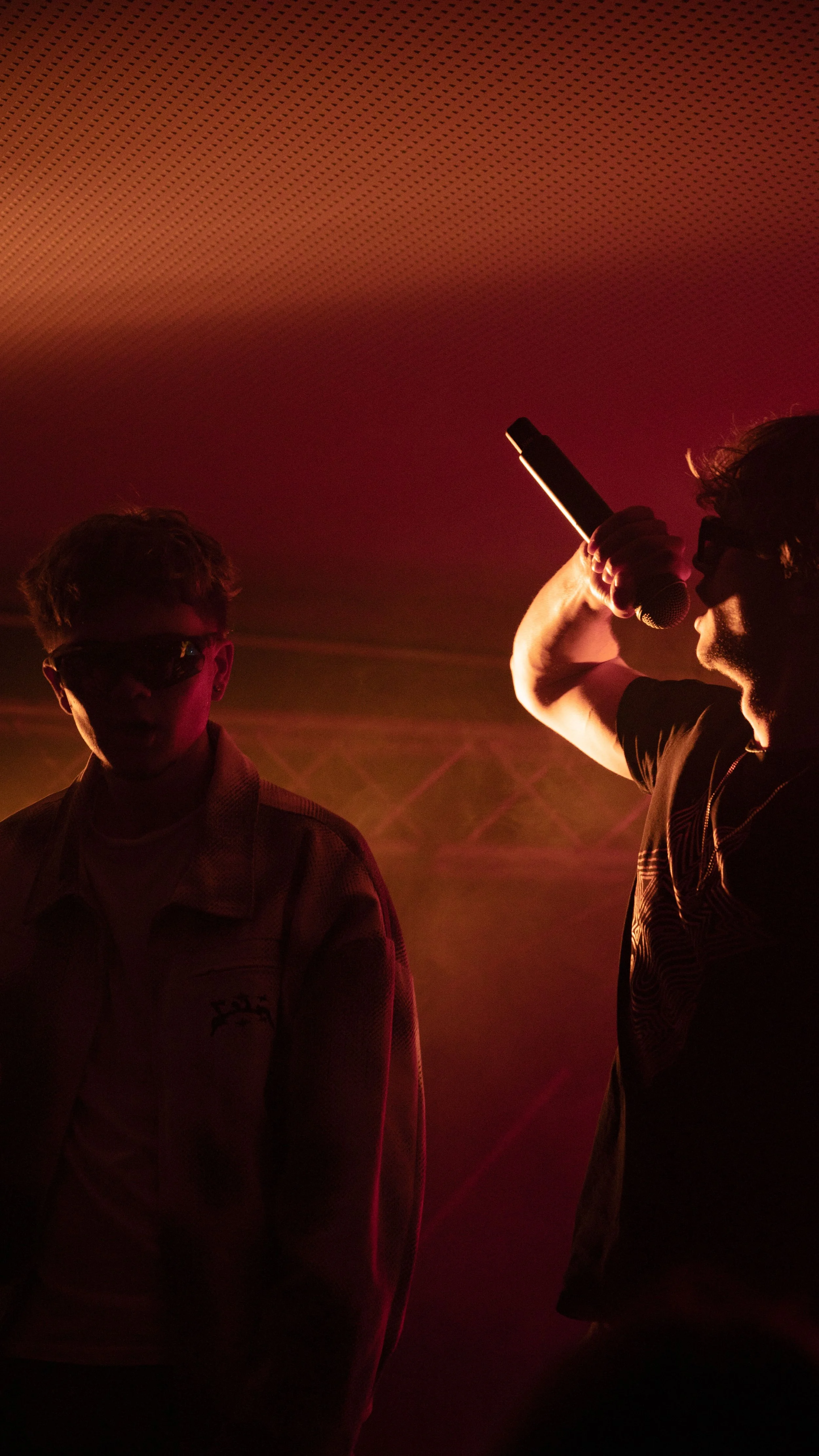 Deux hommes en silhouette, un tenant un microphone, sous un éclairage rouge et orange dans une ambiance de concert ou de scène.