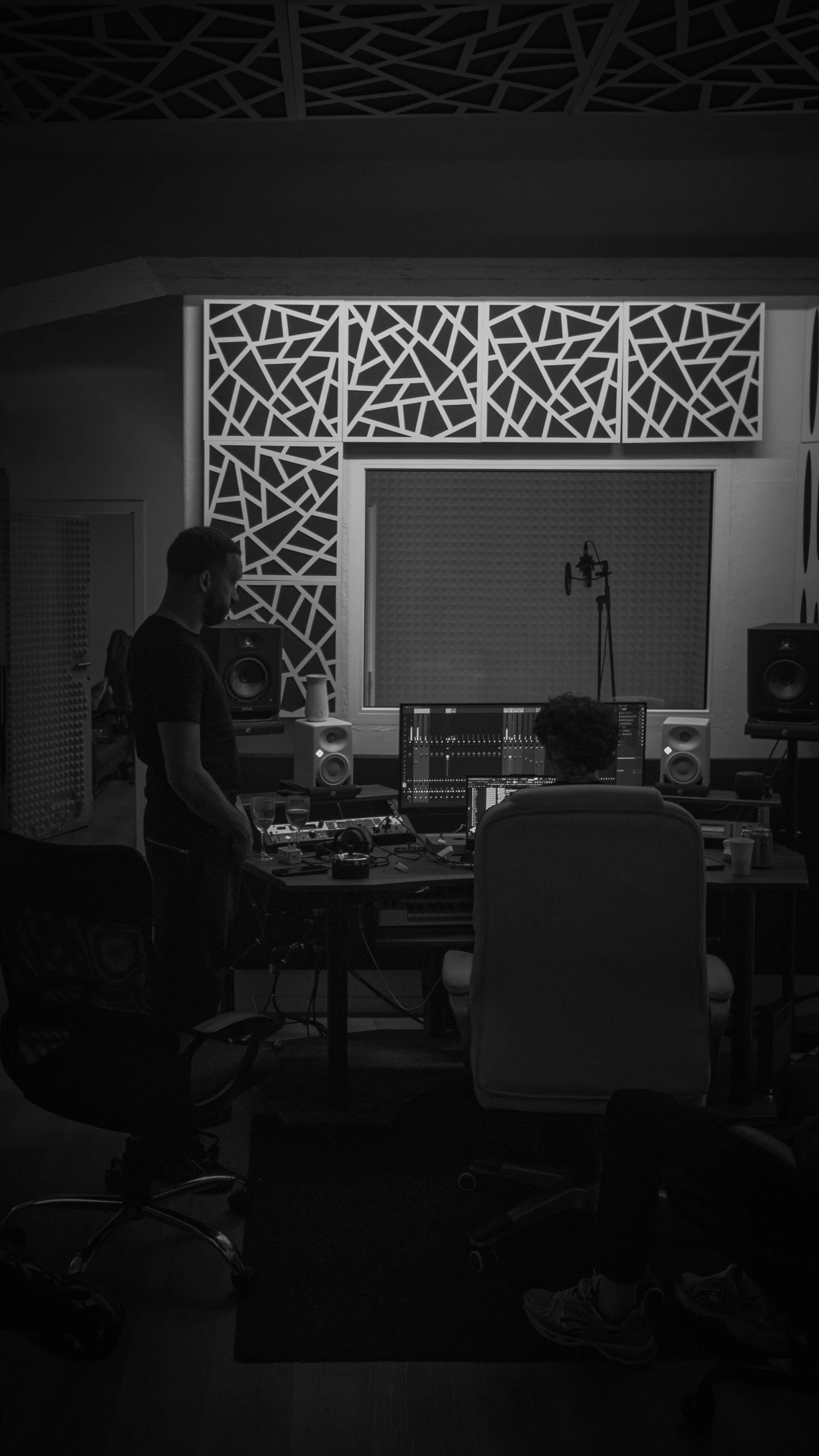 Une salle d'enregistrement audio avec deux personnes, une assise devant un ordinateur et une autre debout à côté, entourées de matériel audio, avec un mur de décoration géométrique et une cabine d'enregistrement dans l'arrière-plan.
