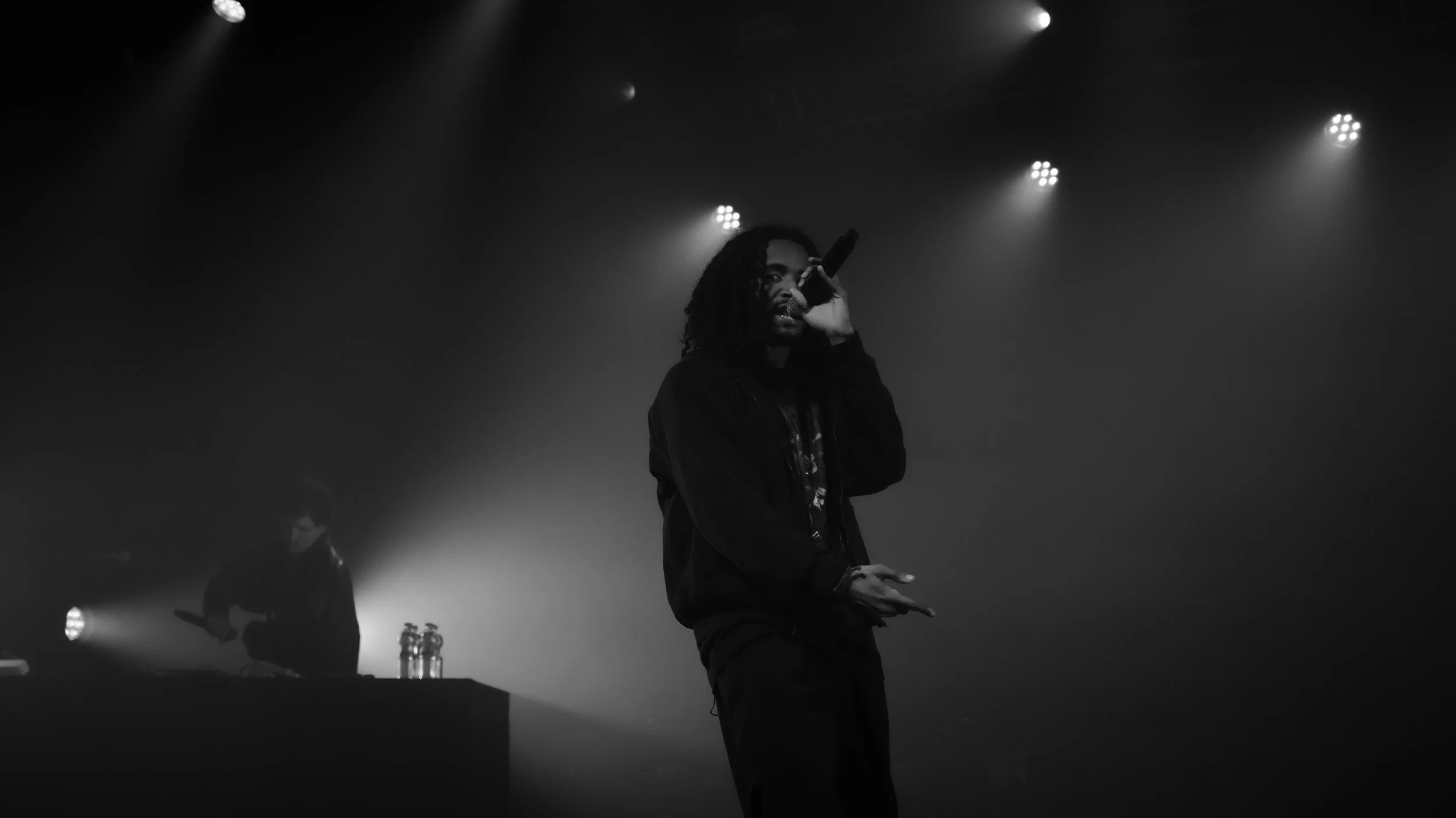 Un artiste de rap ou chanteur en noir et blanc, en train de performer sur scène avec une main tenant un micro, sous des projecteurs lumineux dans l'obscurité.