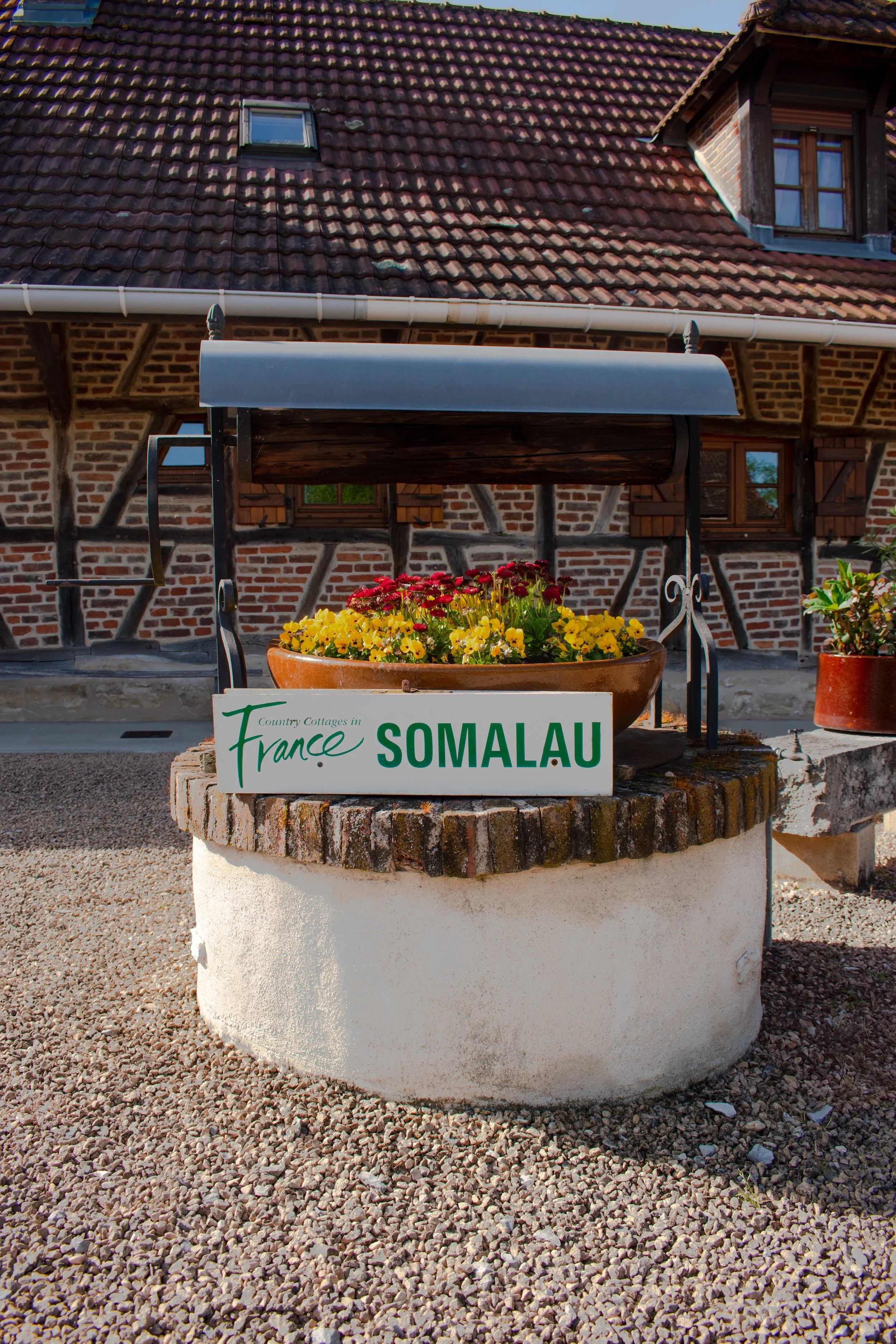 Un panneau indiquant « Somalau » dans un village français, avec une maison traditionnelle en brique et une toiture en tuiles, entouré de plantes en pot et de gravier.